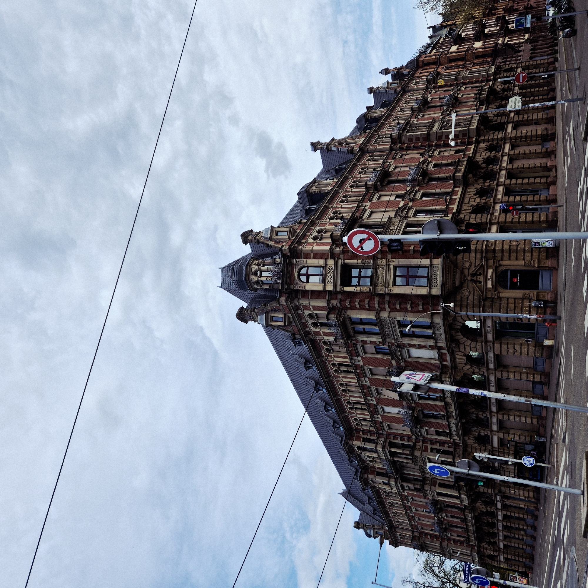 The image captures a view of a tall, ornate building in Strasbourg, France, positioned against a cloudy sky. The building is characterized by its red brick facade and elaborate architectural details, including multiple windows, decorative molding, and a steeply pitched roof with dormers. Traffic signals and street signs are visible along the street, indicating a pedestrian crossing and traffic regulations. The photograph appears to have been taken from a slightly low angle, emphasizing the height of the building. Power lines stretch across the sky in the upper left corner of the frame.