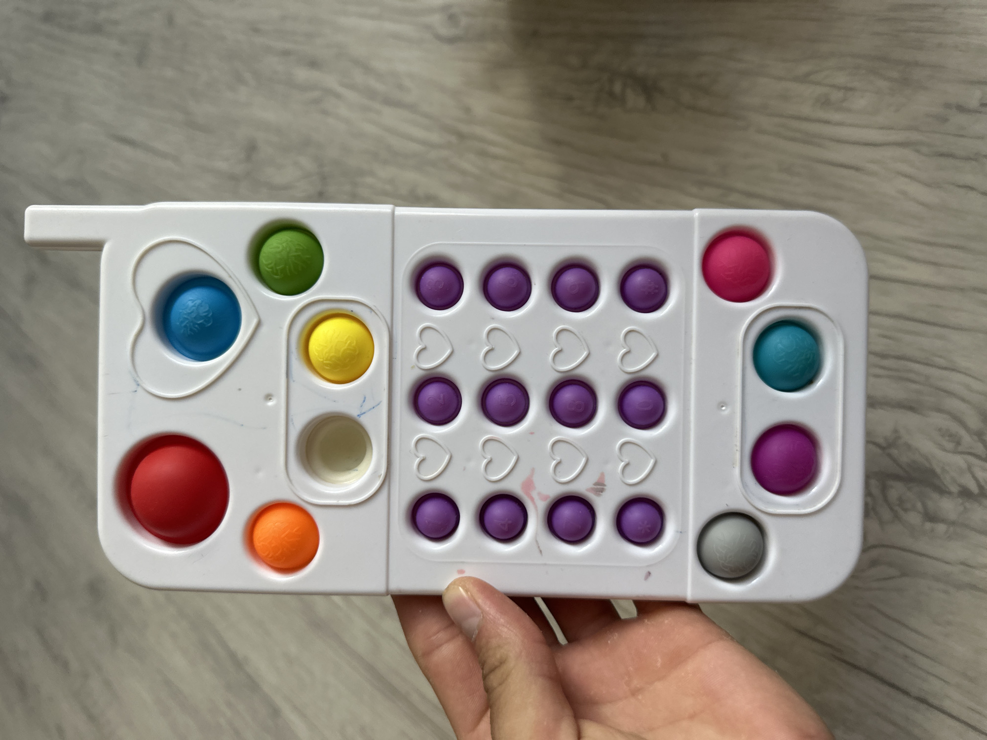 A white, toy cell phone-shaped pop-it toy is lying on a light wood-grained floor. The toy has several colored silicone bubbles. At the top, there's a red bubble, then an orange bubble below it. To the right of the orange bubble is a clear bubble, and to the right of that is a yellow bubble. Above the yellow bubble and to the right of the red one is a blue bubble inside a heart-shaped indent. To the right of the blue bubble is a green bubble. Below these top bubbles is a keypad area with rows of purple bubbles and white heart shapes. The bottom of the toy has three more bubbles: grey on the left, purple in the middle, and teal on the right. A person's hand is visible on the left side of the toy, holding it up slightly. The background is the floor. There is no information provided about the location in the city of Мариинский, Russia, in this image.