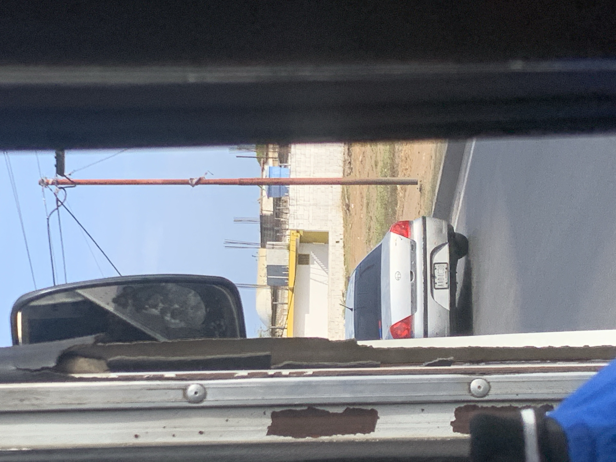 The image captures an outdoor scene from the perspective of a vehicle, likely a bus or large van, looking out its side window. In the immediate foreground, the interior of the viewing vehicle is visible, featuring a dark upper frame, a side mirror reflecting an indistinct scene, and a worn-out window frame with peeling material and rust spots. A portion of blue fabric or garment is visible in the bottom right corner.
In the midground, a silver or light grey hatchback car, possibly a Toyota, is seen on a paved road, facing away from the viewer. Its taillights and rear bumper are clear. A white rectangular license plate is partially visible, showing characters that appear to be "AA01" followed by unreadable symbols, suggesting it might be a Venezuelan plate.
Beyond the road, the background features a construction site. Several unfinished buildings made of concrete blocks are visible, along with some yellow and blue structures. The ground around the construction appears dry and dusty. On the far left, a utility pole with wires and a distinctive long, horizontal orange-red pole extends into the frame. The sky above is clear and bright blue, indicating daytime. The scene suggests a sunny, perhaps warm, climate.