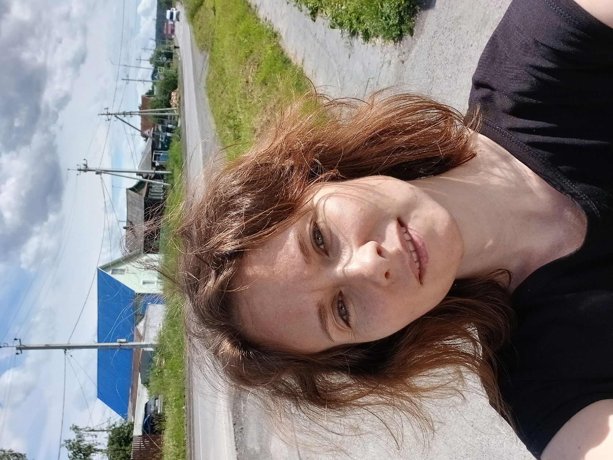 A young woman with reddish-brown hair, freckles, and light-colored eyes poses for a selfie outdoors. She is wearing a black t-shirt. Her hair is windblown. In the background, there is a paved road, a sidewalk, a grassy area, several houses, utility poles, and power lines. The sky is cloudy. The scene appears to be in a rural or suburban area.
