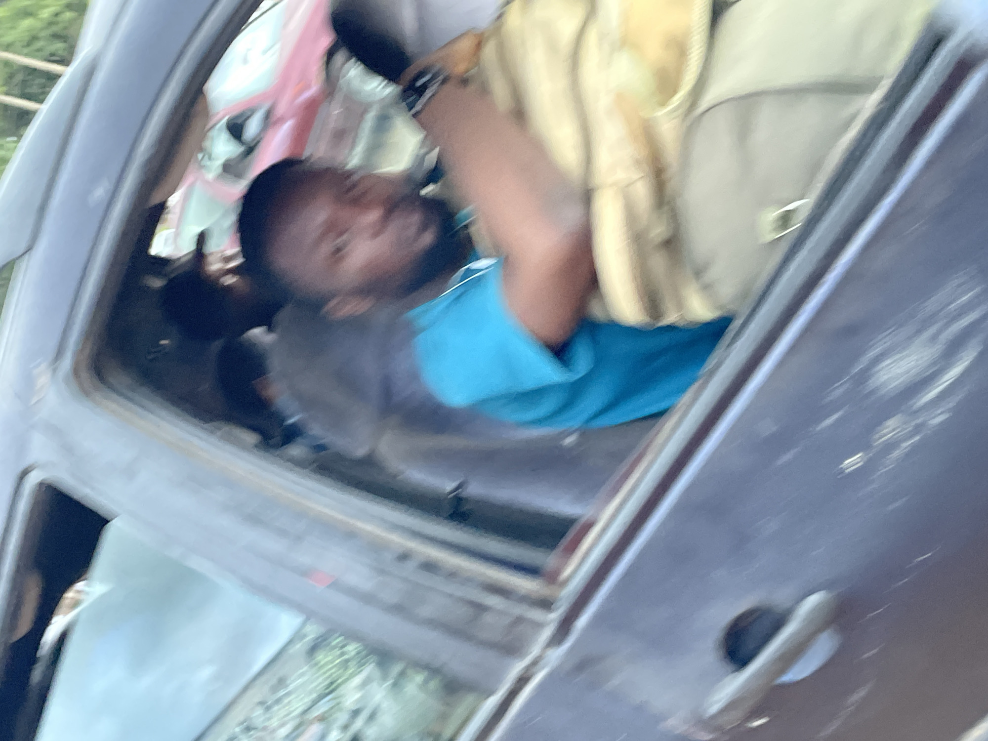 The image is a slightly blurred, close-up interior shot of a dark-colored vehicle, likely taken from outside the driver's side. A man with dark skin, a beard, and short dark hair is visible in the driver's seat, wearing a bright blue t-shirt. He is looking towards the camera with a neutral expression. His right arm is resting on a light brown, possibly canvas bag, which fills a significant portion of the frame. The interior of the car appears worn. Through the window, a red car is visible in the background, suggesting the vehicle is in traffic or parked near other vehicles. The background outside the car is also blurred, but hints of greenery and structures are discernible, indicating an outdoor urban or semi-urban environment. Given the context, this scene could be located in the city of Jos, Nigeria, where such vehicles and traffic conditions are common.