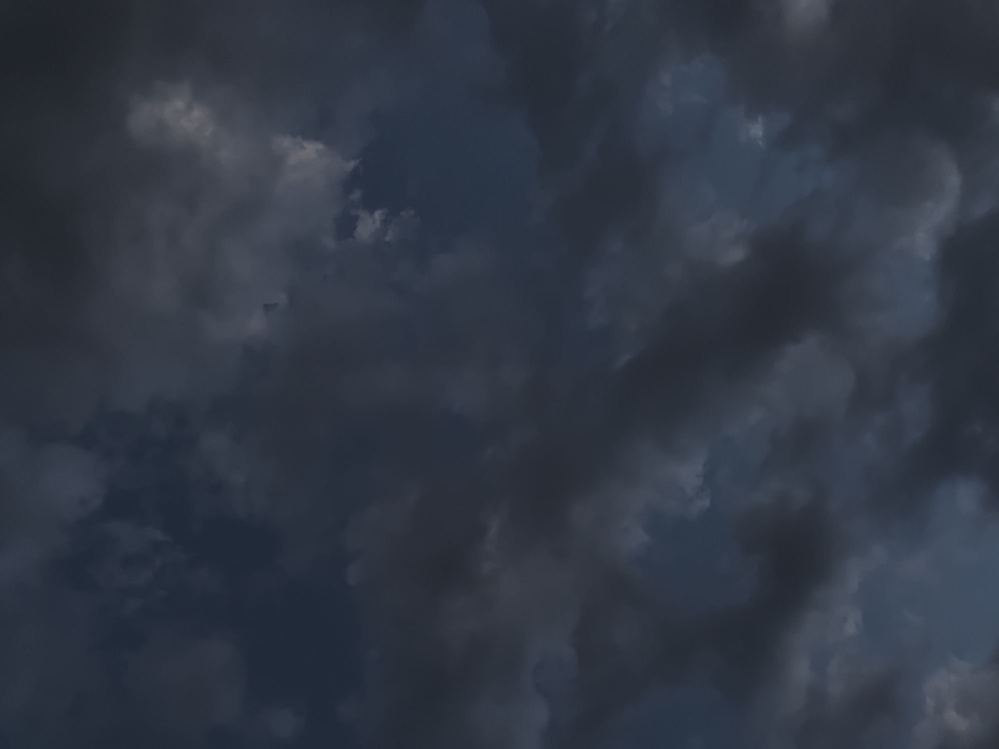 The image displays a close-up view of dark, stormy clouds against a deep blue sky. The clouds are dense and textured, with varying shades of gray and hints of white where light breaks through. The overall impression is one of an impending storm or a sky transitioning from day to night. No specific location in a city is identifiable, as the image is exclusively of the sky.