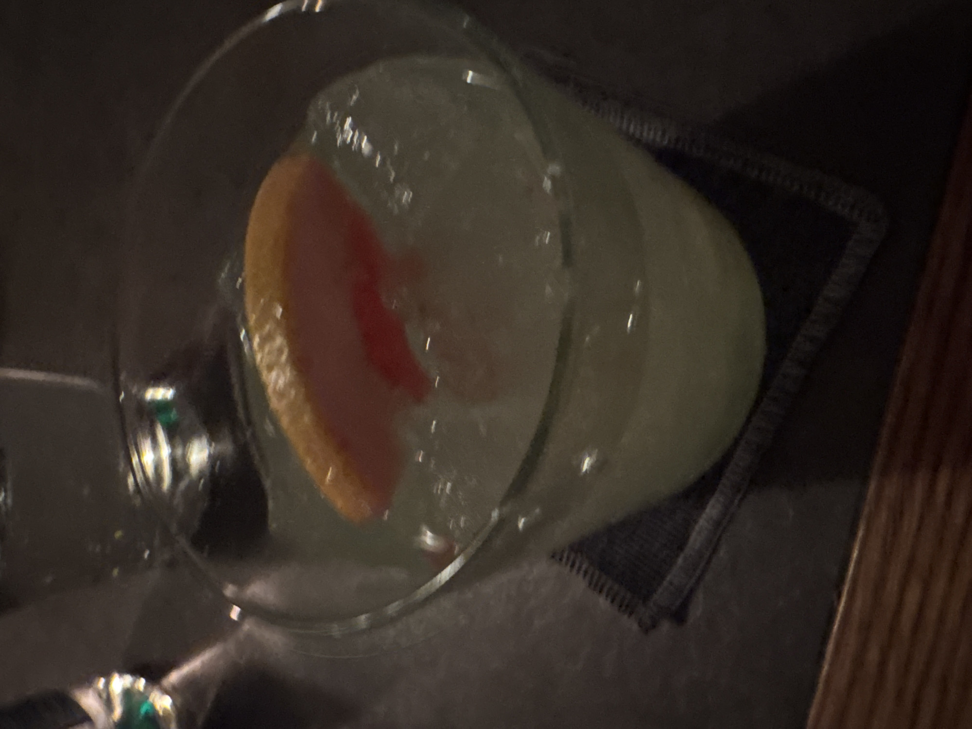 A high-angle shot shows a partially filled clear glass on a dark grey mat. The glass appears to contain ice and a slice of orange or grapefruit. The mat is on a dark surface, possibly a table, with a lighter-colored wooden surface visible in the right corner. The scene is dimly lit. The location is unknown, the image lacks distinctive visual information to identify Plovdiv, Bulgaria.