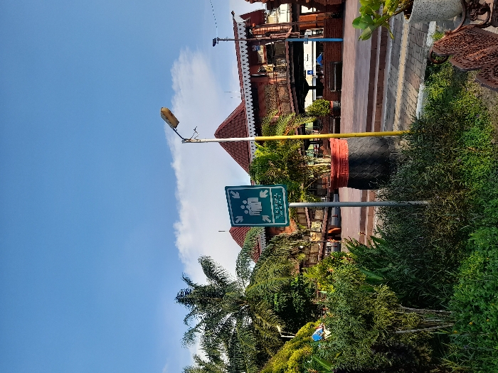 The scene is located at -7.464 latitude and 112.432 longitude in Mojokerto, Indonesia. A green sign with the word "TURUN KEREPA" and an image of people walking in different directions is attached to a wooden pole in front of a red-roofed building. The building appears to be a station or a terminal with multiple entry/exit points, including a shaded walkway with an open-air area and a brick platform. Green bushes are in front of the building and on both sides of the wooden pole. A streetlight is seen on the right side of the pole, and a potted tree is in front of the building. A couple of benches are on the ground in the foreground of the image. The sky is blue and cloudless.