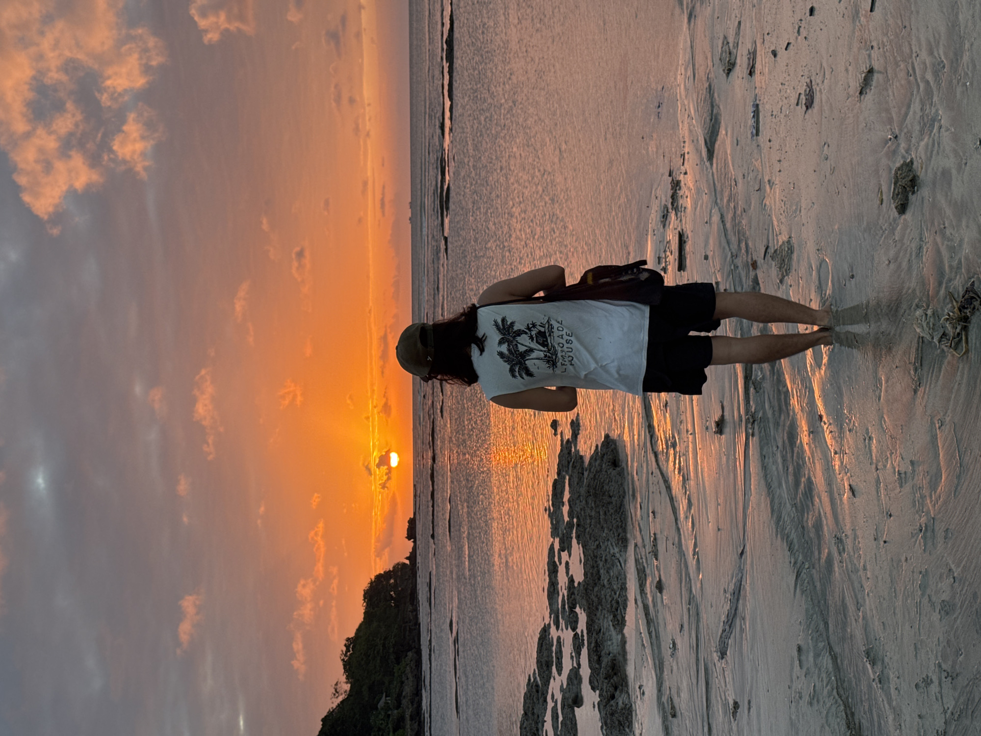 A person stands on a sandy beach in Kuta Selatan, Indonesia, facing the ocean as the sun sets. The sky is ablaze with orange and pink hues, with clouds scattered across it. The water is calm, reflecting the colors of the sunset. The person, seen from behind, is wearing a white tank top with a palm tree graphic, black shorts, and a dark baseball cap. They have a bag slung over their shoulder and are standing in shallow water, with their feet partially submerged. The foreground shows wet sand with ripples and scattered rocks and debris. In the distance, a lush green hillside with trees and a small structure is visible on the left, and the horizon shows a clear line between the ocean and the sky, with faint silhouettes of ships.