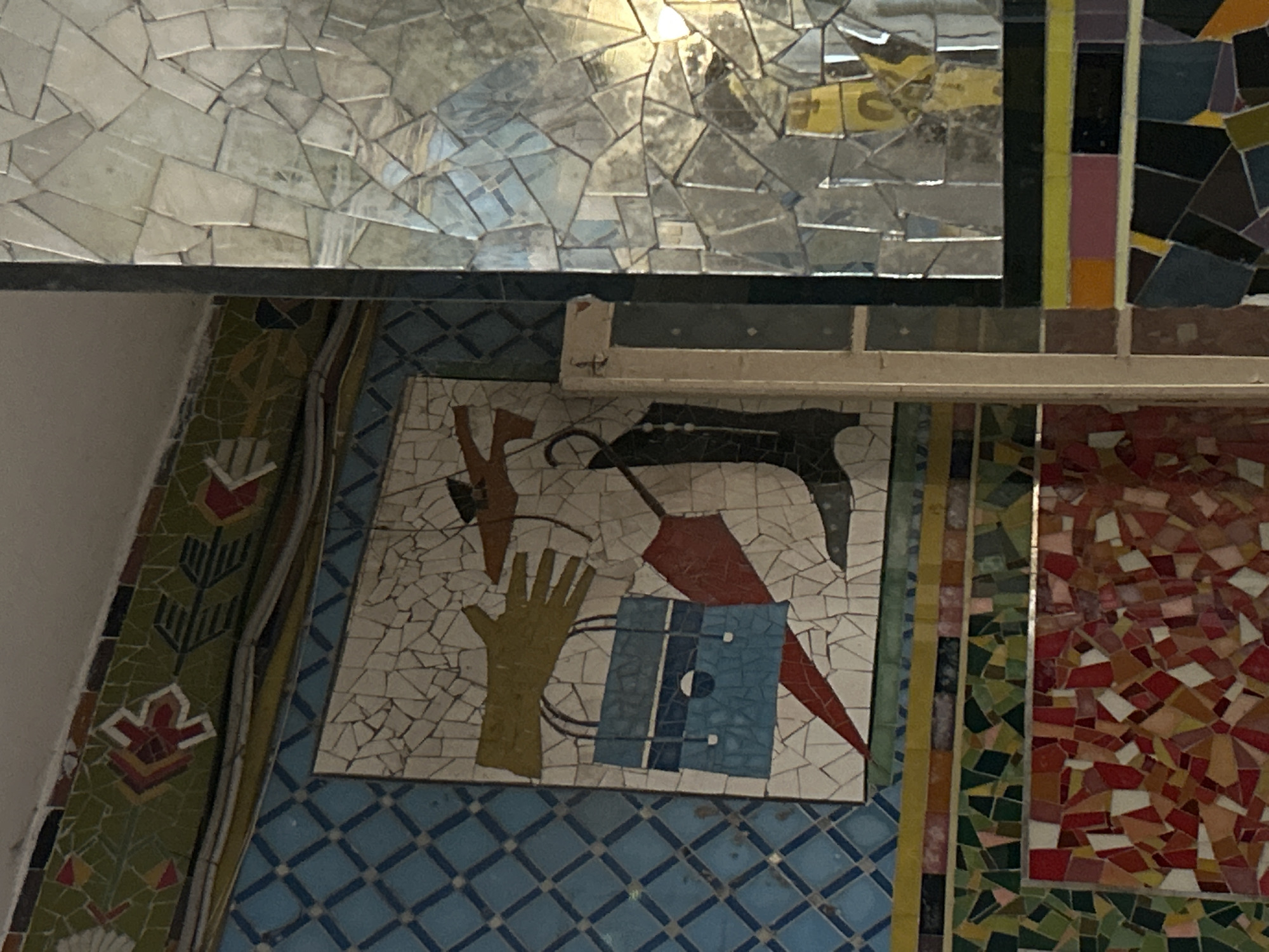 The image shows a wall adorned with colorful mosaics. In the center, a mosaic depicts a yellow gloved hand reaching towards a brown object, possibly a bird. Below this, a blue handbag with white and dark blue stripes is shown, with a red umbrella to its right and a black boot to its left. The wall is decorated with a pattern of blue diamond-shaped tiles to the left of the central mosaic. Above this, there is a decorative border with green and red mosaic patterns. To the right of the central mosaic, a column covered in mirrored mosaic tiles reflects the surroundings. A section of the wall below the central mosaic features a vibrant mosaic of red, pink, and yellow tesserae. The overall scene suggests an interior decorated with artistic mosaic work. The location is identified as the Grigore Antipa National Museum of Natural History in Slobozia, Moldova.