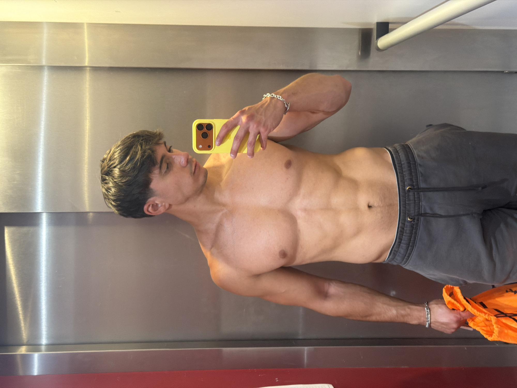 A shirtless man with dark, styled hair takes a selfie in an elevator. He is wearing grey sweatpants and a silver bracelet on his left wrist. In his left hand, he is holding an orange drawstring bag. The elevator appears to be in Sofia, Bulgaria, judging by the presence of a railing often found in Bulgarian public transport. The elevator walls are metallic, with a red panel visible on the left side. The lighting is bright, casting shadows on the man's physique.