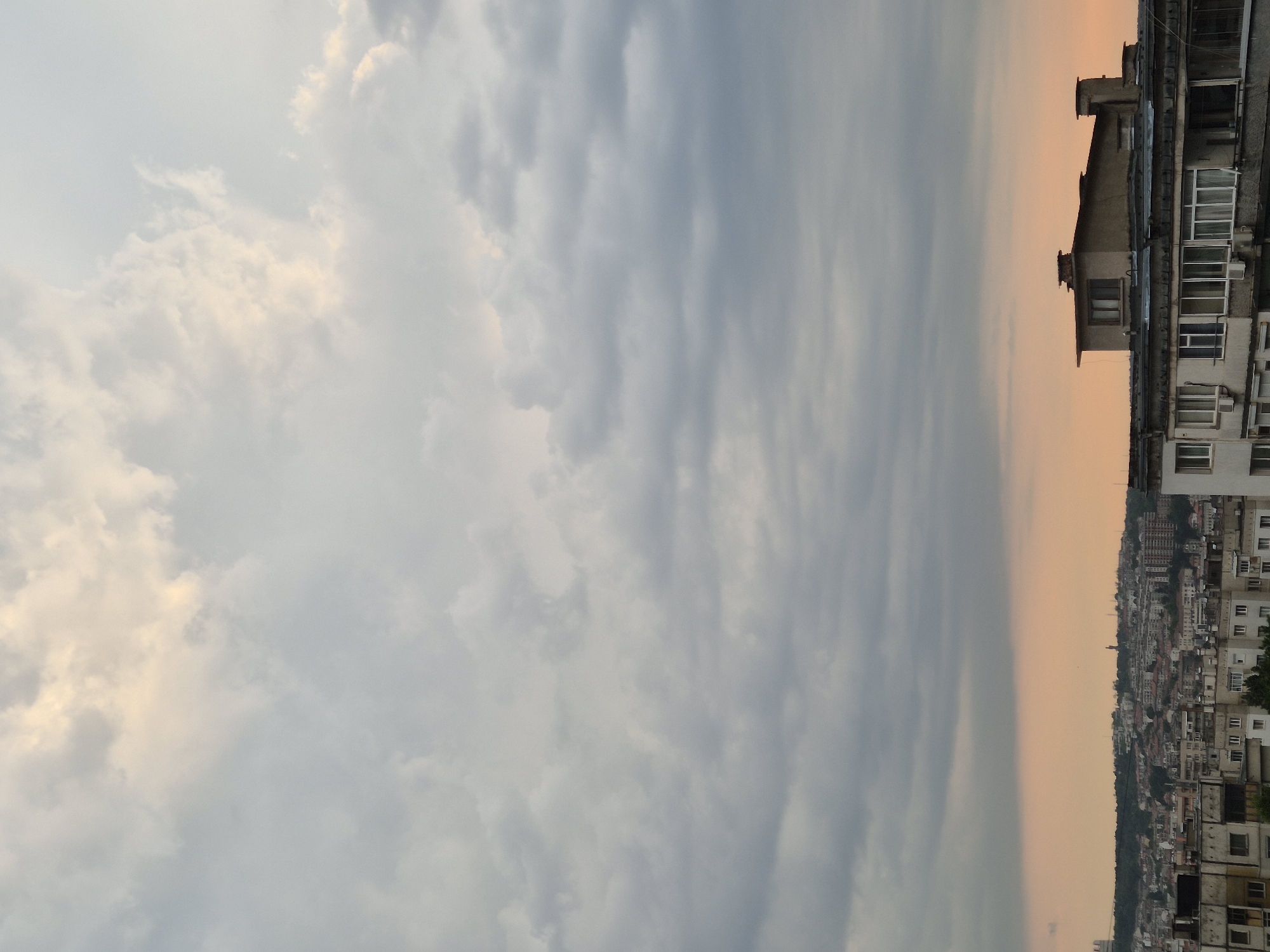 The image depicts a city skyline under a cloudy sky at dusk. The sky transitions from a bright, pale yellow with fluffy clouds on the left to a muted, grayish blue with streaky clouds on the right, suggesting the setting sun is on the horizon. The cityscape is visible on the right, characterized by a dense collection of buildings with flat roofs and many windows. The architecture appears to be of a uniform style, possibly apartment buildings. The overall impression is one of urban sprawl under a dramatic sky. The lighting suggests it is either early morning or late evening. There is no visible text in the image.