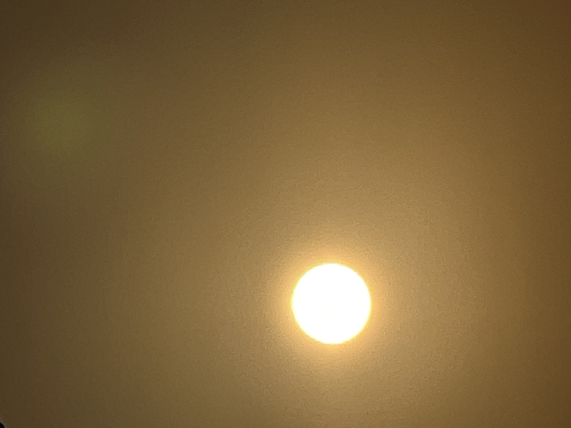A bright white, circular sun is visible in the left-center of the frame, partially obscured by atmospheric haze, which tints the sky a uniform yellowish-brown. A faint, slightly green circular lens flare is also present in the upper right quadrant of the image. The overall scene suggests a hazy or overcast day, possibly during sunrise or sunset, as the sun's intensity is diffused. The setting is a broad, indistinct sky, with no discernible landmarks or structures to indicate a specific location within Kuta, Indonesia, or any other city. The image captures a natural phenomenon rather than a specific urban scene.