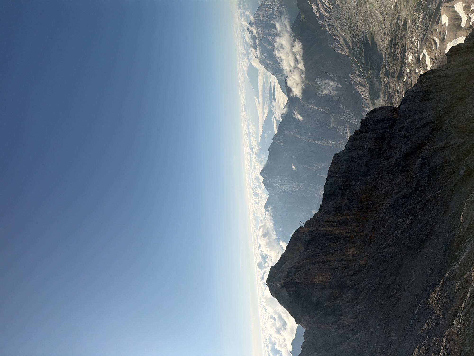 The image depicts a vast mountain landscape with a clear blue sky dominating the upper portion. Below the sky, a layer of fluffy white clouds stretches across the horizon, partially obscuring a range of rugged, blue-toned mountains. In the foreground, a sharp, dark mountain peak juts out, with rocky scree slopes descending into the valley. A small, dark silhouette of a person stands on the edge of this prominent peak, appearing to be observing the view. Further into the distance, more mountain peaks and valleys are visible, with patches of snow or ice on some of the lower slopes. A thin, dark pole-like structure is visible on the right side of the foreground peak. The scene is likely captured from a high vantage point, offering a panoramic view of the alpine environment. This could be the Zugspitze, Germany's highest peak, located near Grainau.