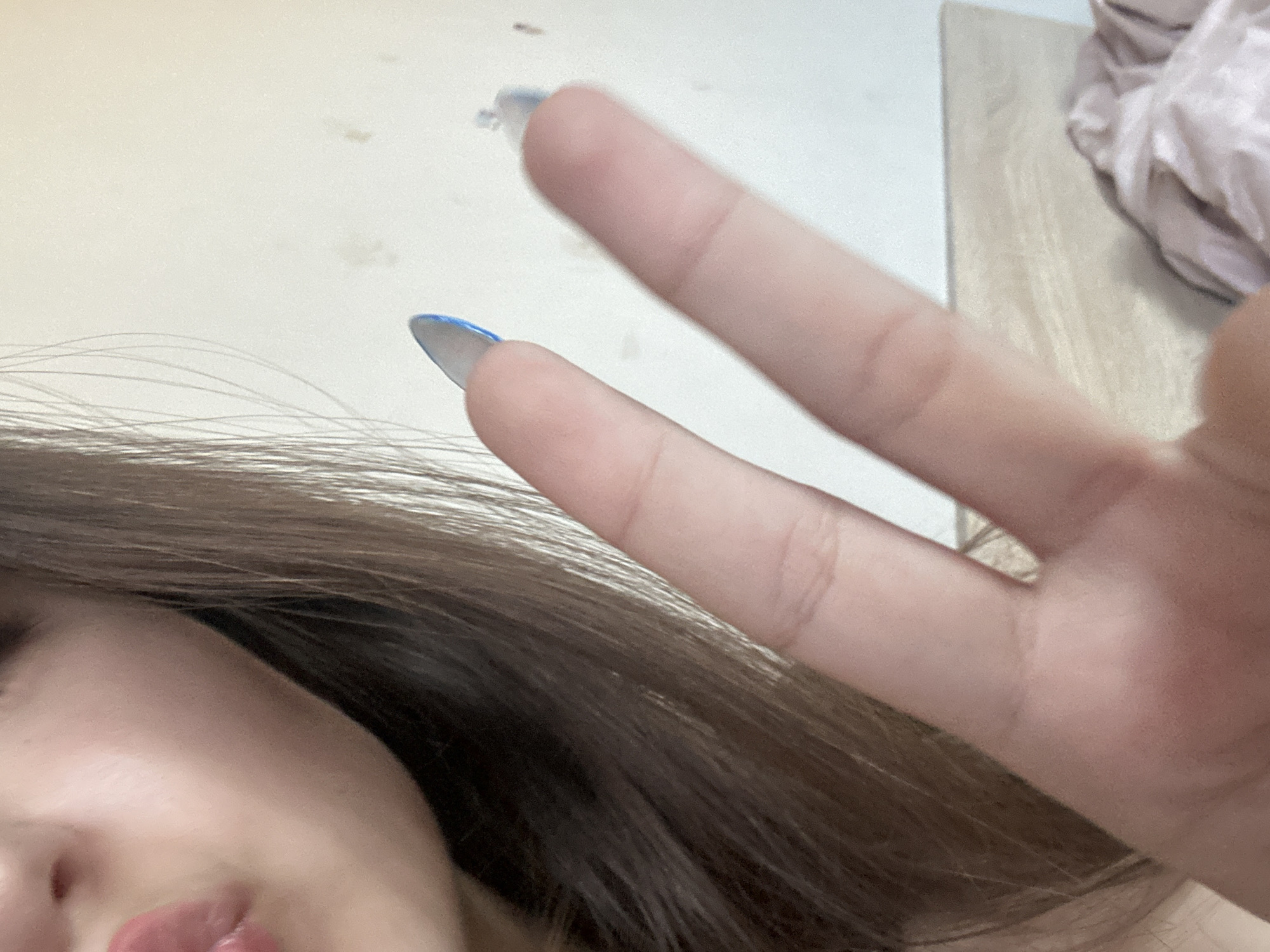 The image shows a close-up, selfie-style shot of a person's face and hand. The person has long, brown hair that frames their face. A portion of their left cheek and lips are visible, with what appears to be light pink lipstick. The person's right hand is in the foreground, with the index and middle fingers extended in a peace sign. The nails are painted with blue polish. The background is a plain, light-colored wall, with some indistinct objects or textures visible. The lighting suggests an indoor setting, possibly during the day given the brightness. No text is visible in the image.