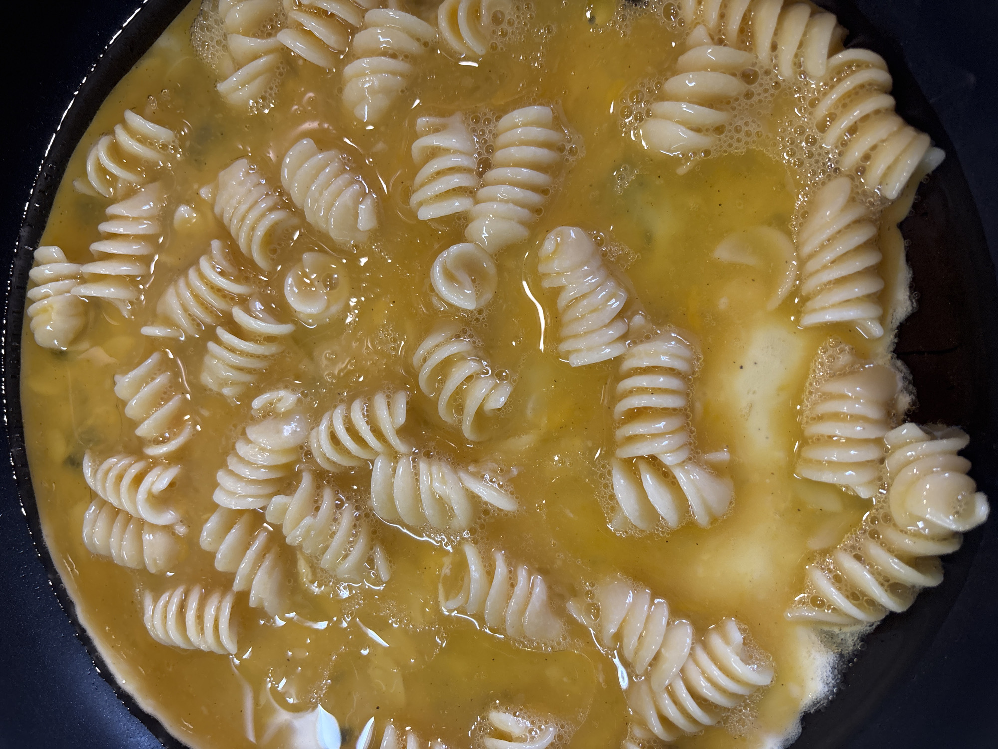 The image shows a close-up, overhead view of fusilli pasta being cooked in a pan with what appears to be scrambled eggs. The pasta is a pale yellow-white, and the egg mixture is a vibrant yellow, with some areas appearing slightly darker due to cooking. Bubbles are visible in the egg mixture, indicating it is hot and actively cooking. The pasta is distributed throughout the pan, submerged in the egg. The pan itself is black and has a slightly raised edge, suggesting it is a non-stick skillet. There are no people visible in the image. The scene depicts food preparation. There is no information available to determine the location in the city of Bila Tserkva, Ukraine.