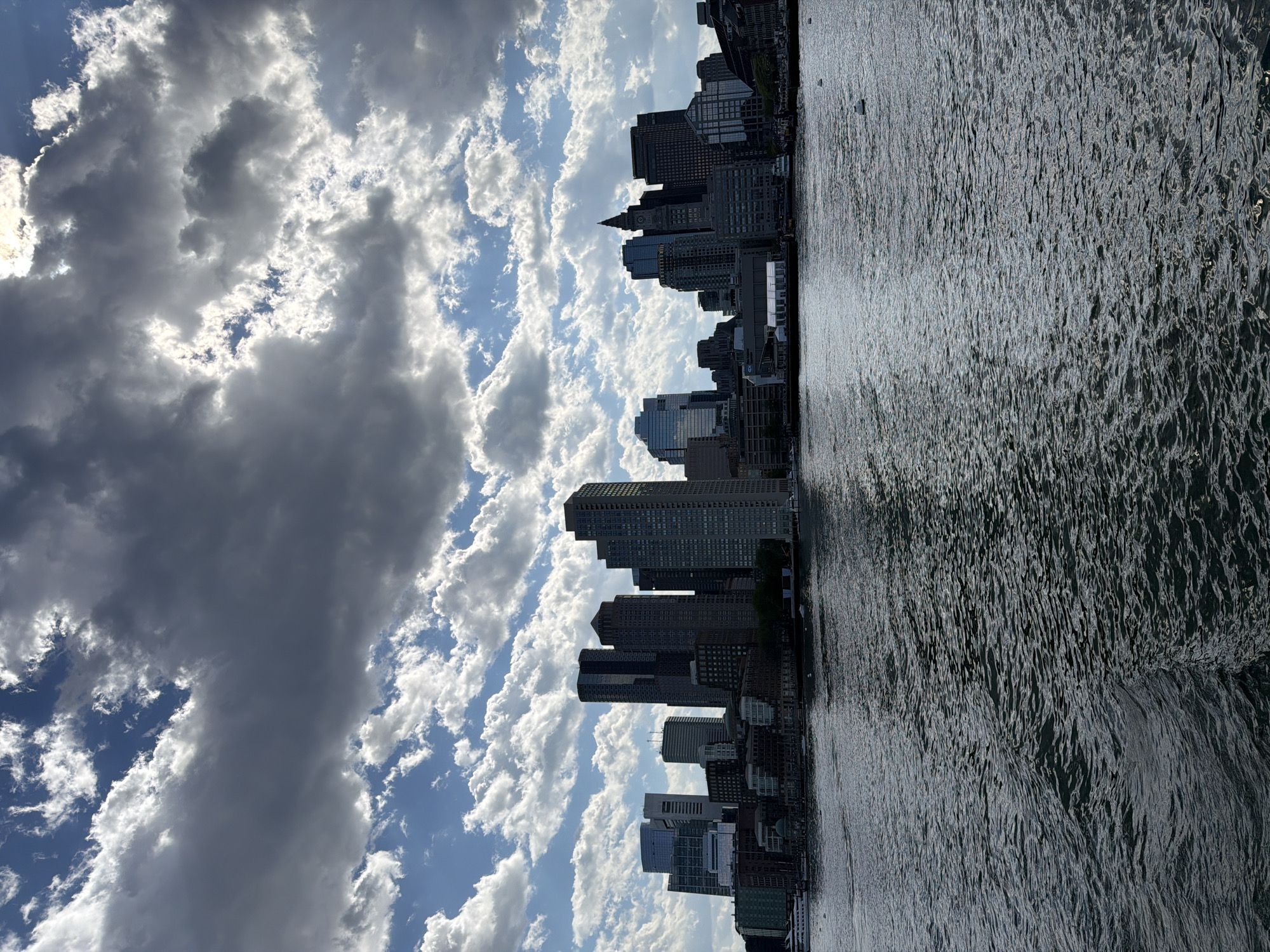 The image shows the Boston skyline from across the harbor. The buildings are mainly tall skyscrapers, silhouetted against a partly cloudy sky. The water in the foreground is slightly choppy, reflecting the sunlight. The location appears to be the downtown area of Boston. There are no people visible in the image.