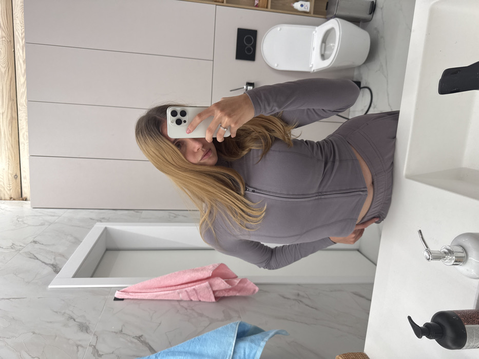 A woman in a grey zip-up athletic top and matching pants takes a selfie in a bathroom. The bathroom has white cabinetry, a white marble floor with a blue and pink towel in the corner. A white toilet is visible in the background, along with a white sink with soap dispensers in the foreground. The image is rotated.