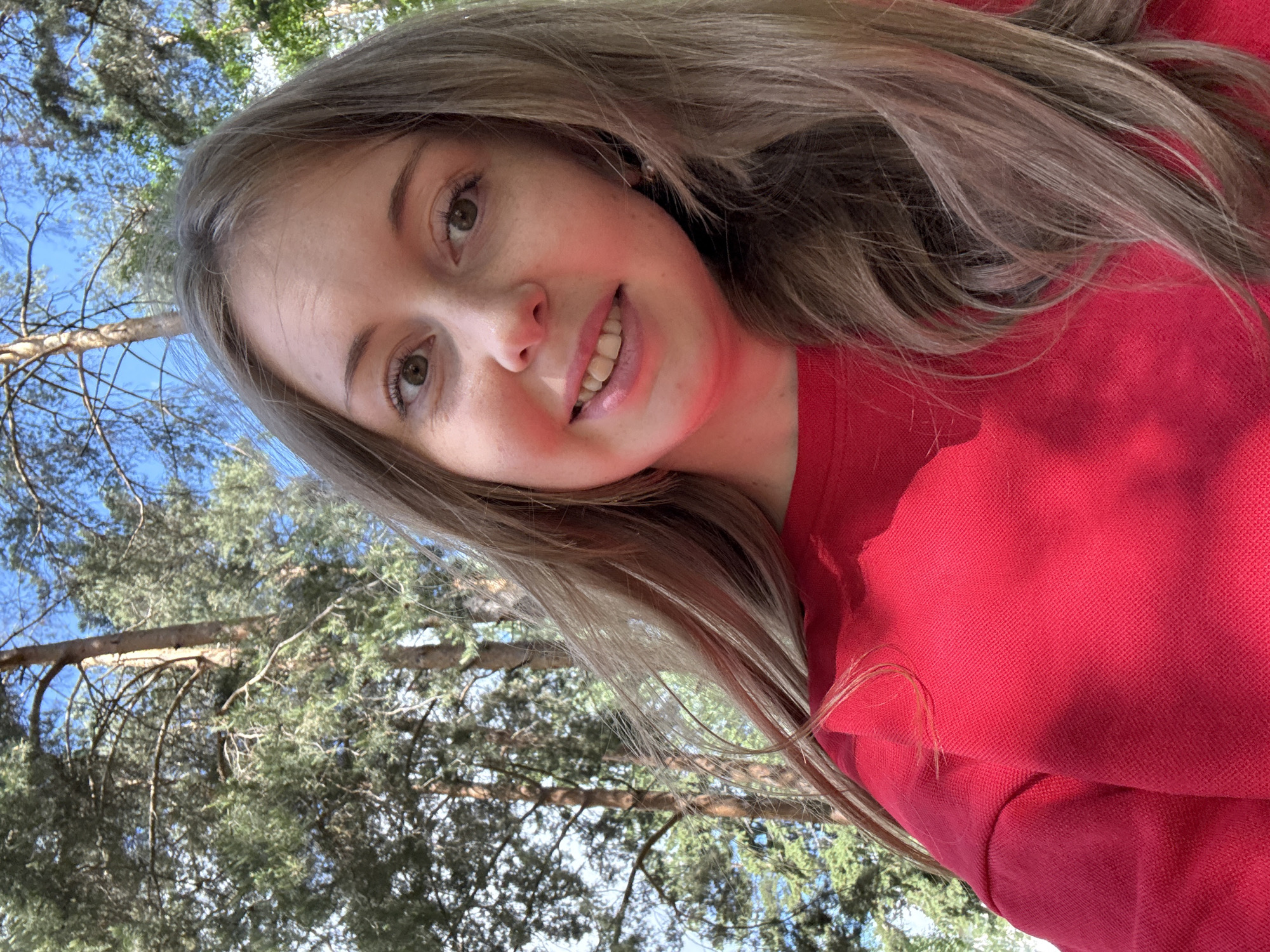 A woman with light brown hair is wearing a red sweater and smiling at the camera. She is outdoors, and in the background are trees and a blue sky. The exact location is Unknown, Unknown.