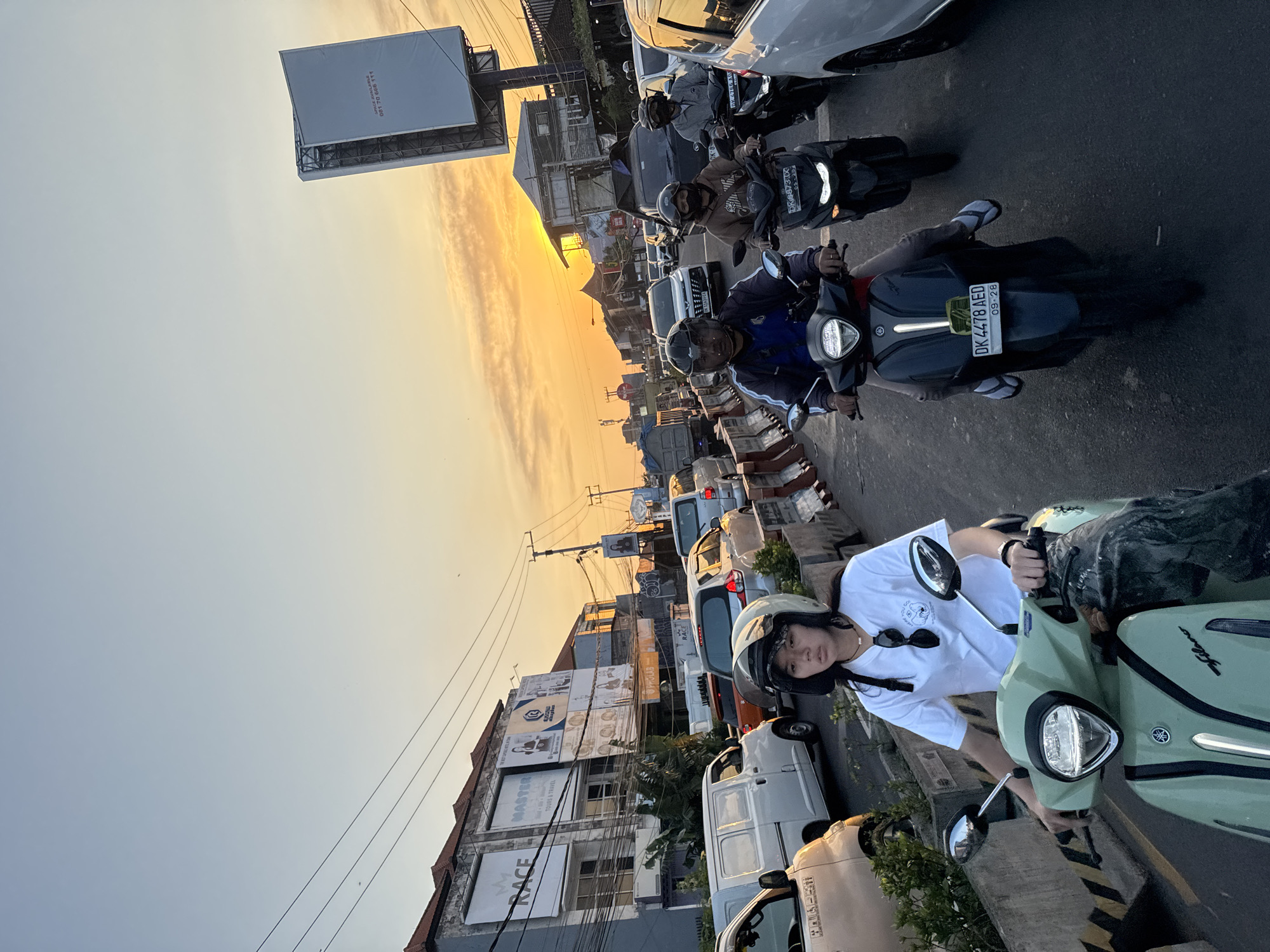 The image depicts a busy street in Denpasar, Indonesia, during a sunset. The sky is illuminated by the warm hues of the setting sun, casting a soft glow over the scene. The street is congested with vehicles, including cars, vans, and numerous motorcycles. Several people are seen riding motorcycles, many wearing helmets, indicating a typical traffic situation in the city. Buildings line the street, some featuring advertisements and signs, suggesting a commercial area. Power lines and poles are visible overhead, typical of urban infrastructure. The overall impression is one of a vibrant city caught in the hustle and bustle of rush hour traffic, bathed in the beautiful light of a tropical sunset.