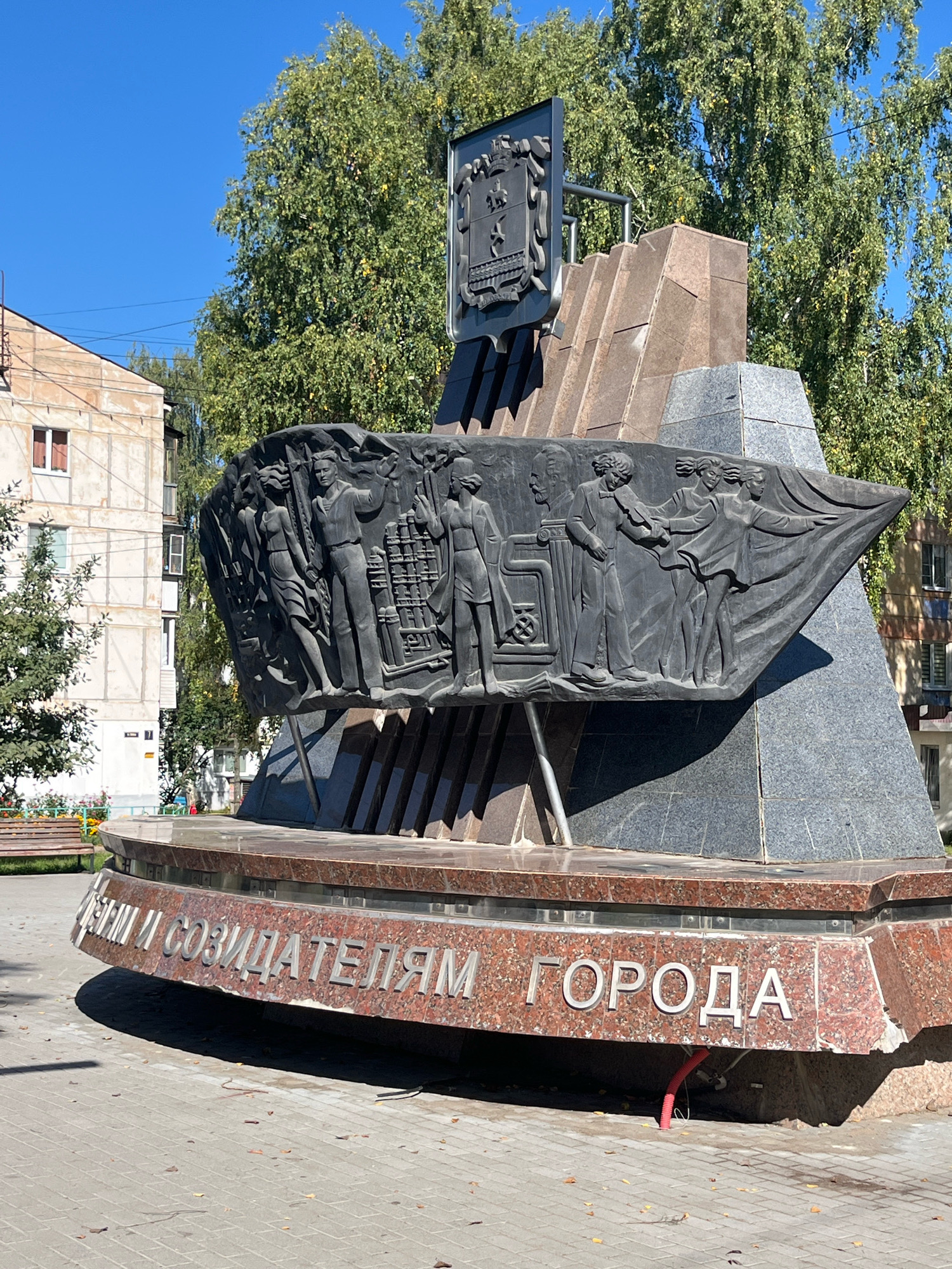 The image shows a monument in the city of Chaikovsky, Russia. The monument features a large, dark bas-relief depicting several figures in historical attire. The figures appear to be engaged in some form of collective activity, possibly related to construction or development. Above the bas-relief, there is a tiered structure made of granite and stone, topped with a coat of arms. The base of the monument is adorned with metal lettering in Russian, which translates to "To the founders and creators of the city." The monument is situated in an outdoor paved area, possibly a public square or park, with trees and buildings visible in the background. The sky is clear and blue, suggesting a sunny day.