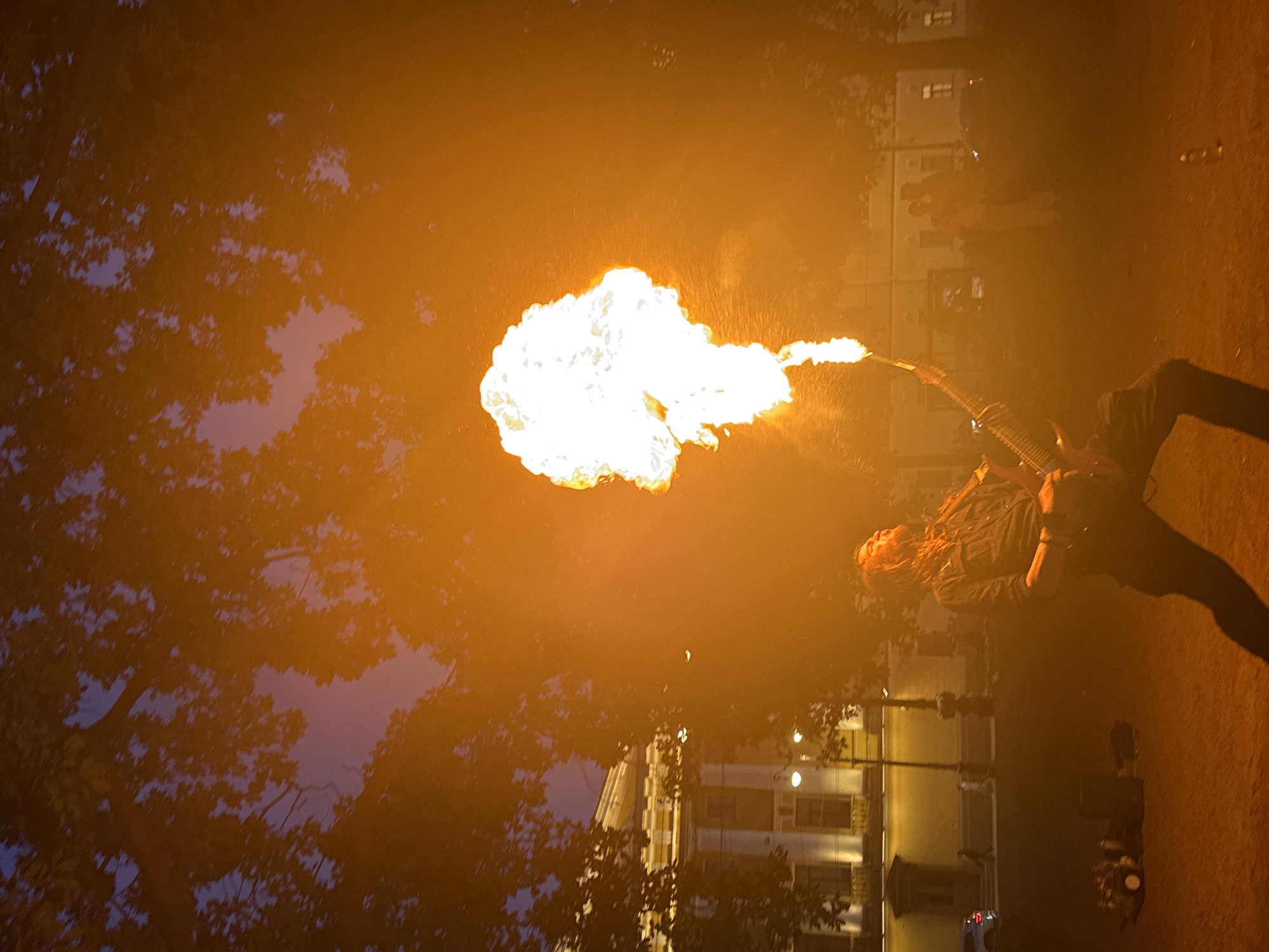 The image depicts an outdoor fire performance occurring at twilight or night in Saint Petersburg, Russia. In the lower-center-left, a person, appearing male with long, light-colored hair and a beard, is performing with a custom instrument resembling an electric guitar. A large, voluminous, white-yellow flame, estimated to be several meters high, erupts upwards from the instrument's headstock/neck area, forming a mushroom-like cloud. The performer is clad in a dark collared shirt and dark trousers, with his head tilted back and mouth slightly open, focused on the ascending flame.
The immediate ground surface is unpaved, reddish-brown earth or gravel. To the performer's left, on the ground, are various pieces of equipment, including what appear to be portable speakers or amplifiers and potentially a small drum or percussion setup. In the midground, to the right of the performer, several indistinct human silhouettes are visible, possibly spectators.
The background on the left reveals a multi-story building featuring classical architectural elements such as columns and cornices, characteristic of Saint Petersburg. Its facade and windows are illuminated by artificial lighting. A street lamp is visible on the sidewalk in front of this building, with a parked vehicle and faint figures of pedestrians on the street. The upper portion of the image is dominated by the dark, silhouetted foliage of numerous trees, obscuring parts of the structures and contrasting against a dim twilight sky. The sky transitions from a darker blue/purple hue at the top to lighter orange and pink tones closer to the horizon, largely influenced by the intense light from the fire performance.