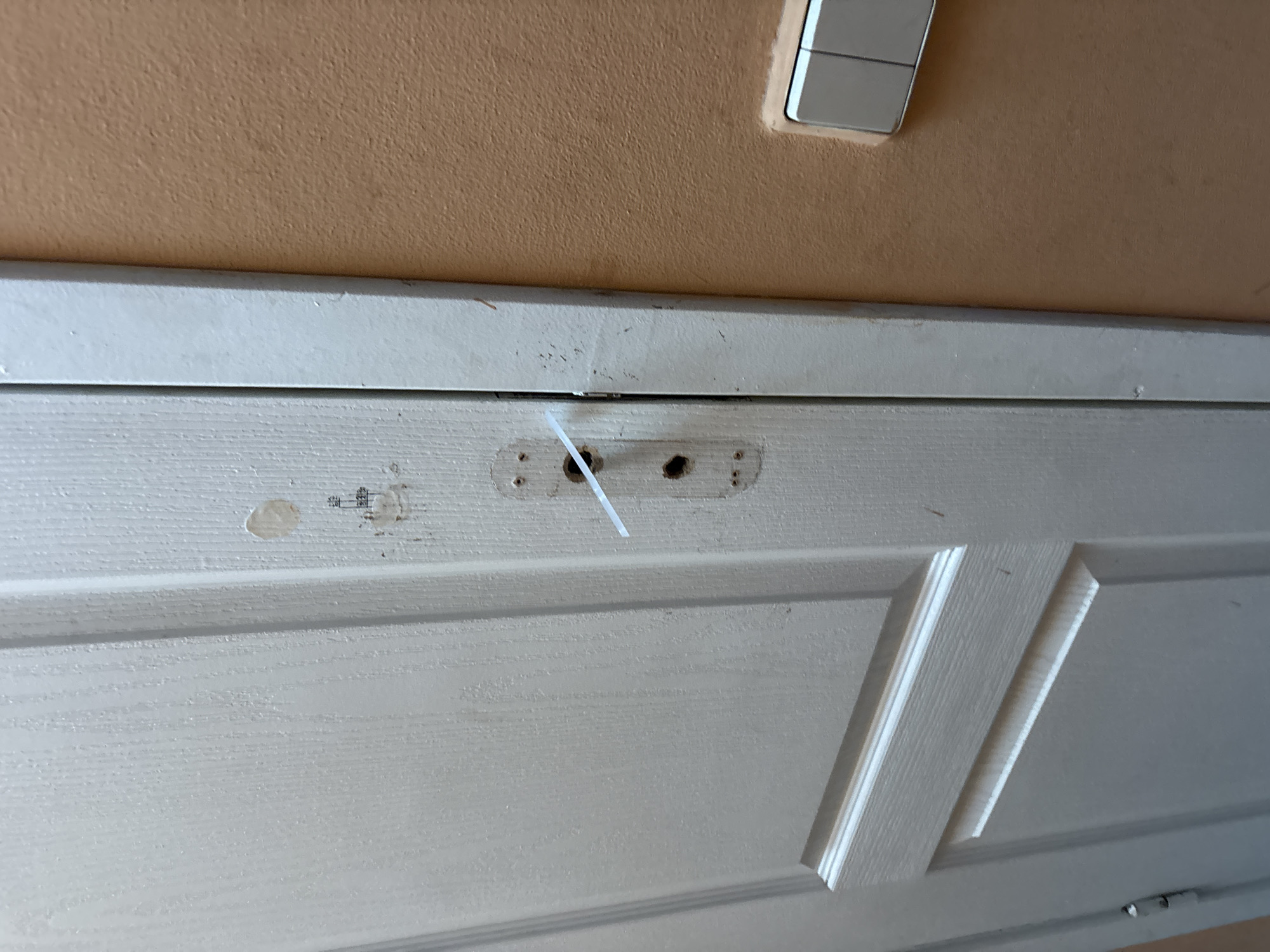 The image shows a close-up view of a white wooden door. The door has a panel design with raised sections. Above the door, a textured peach-colored wall is visible, with a light switch plate in the upper right corner. The light switch has two rectangular buttons.
In the center of the door, there is a small, metal plate with two visible screw holes and what appear to be two keyholes or mounting points. A white zip tie is threaded through one of these openings. To the left of the plate, there's a circular light-colored stain or mark on the door. To the right of the plate, there are some faint markings that might be text, but they are illegible.
The lighting in the image suggests it was taken indoors, possibly during the day, given the illumination and shadows. The overall scene is domestic and utilitarian, with no people or specific activities evident. The focus is on the details of the door and its hardware.