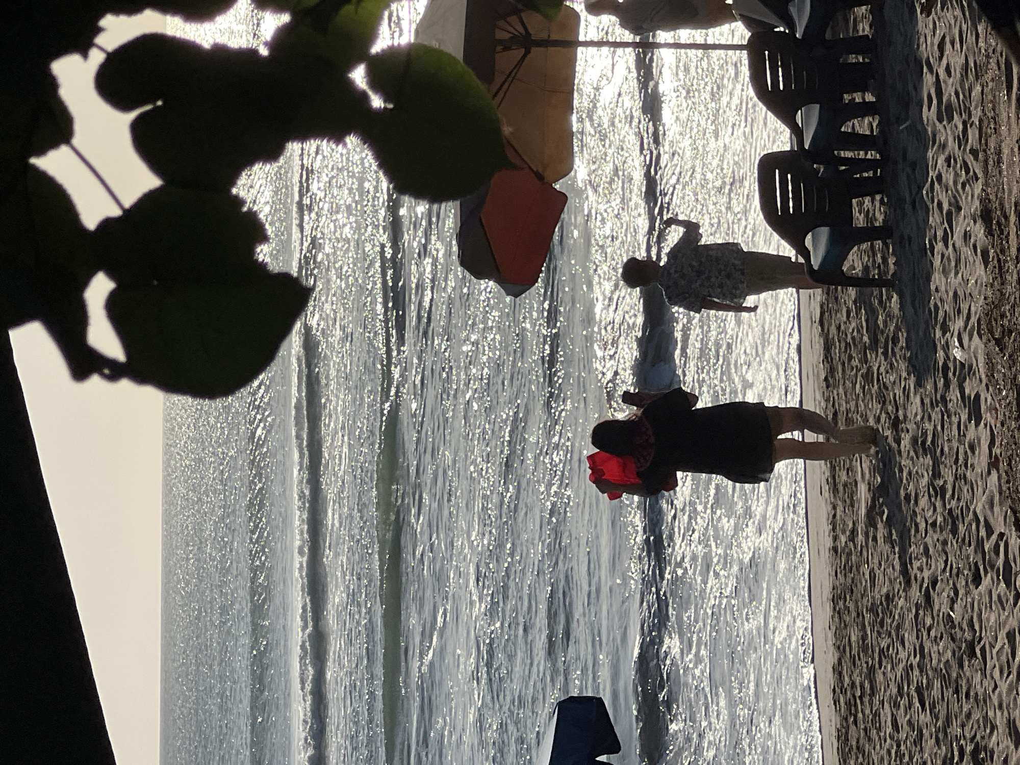In Kuta, Indonesia, a sun-drenched beach scene unfolds as two individuals walk near the water's edge. A woman, viewed from the back, carries a bright red bucket and appears to be walking away from the ocean. Beside her, a person in a patterned short-sleeved shirt walks towards the water. In the background, the sun glints off the ocean waves, creating a sparkling effect. Beach chairs and umbrellas are set up on the sand, suggesting a leisurely atmosphere. Foliage casts shadows on the scene, adding depth and framing the view of the sea.