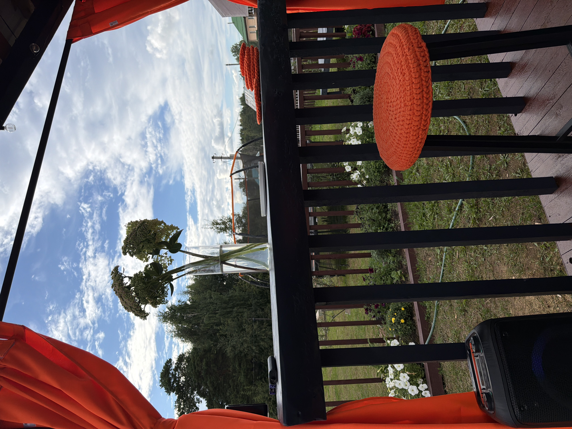 The image depicts an outdoor seating area on a wooden deck, likely a balcony or patio, overlooking a garden and trees. A black railing with vertical supports runs across the foreground. On the deck, a black stool with a bright orange crocheted seat is prominently placed. A black portable speaker is situated on the deck near the railing on the left side.
On a black tabletop or counter behind the railing, a tall glass vase holds several sprigs of greenery with small, pale flowers. A black electronic device, possibly a vape or small remote, rests on the tabletop. To the right of the vase, a coiled orange crocheted item, possibly a placemat or coaster, is placed on the tabletop.
The background reveals a lush green garden with various flowering plants, including white petunias and some darker purple and yellow flowers. Beyond the garden, a line of trees forms a dense green backdrop, with a few houses and what appears to be a trampoline visible in the distance. The sky above is a vibrant blue, dotted with scattered white, puffy clouds. Orange curtains are draped on either side of the frame, framing the view. The overall impression is a relaxed, outdoor recreational space, possibly in a residential area in Chaikovsky, Russia, given the mention of the city.