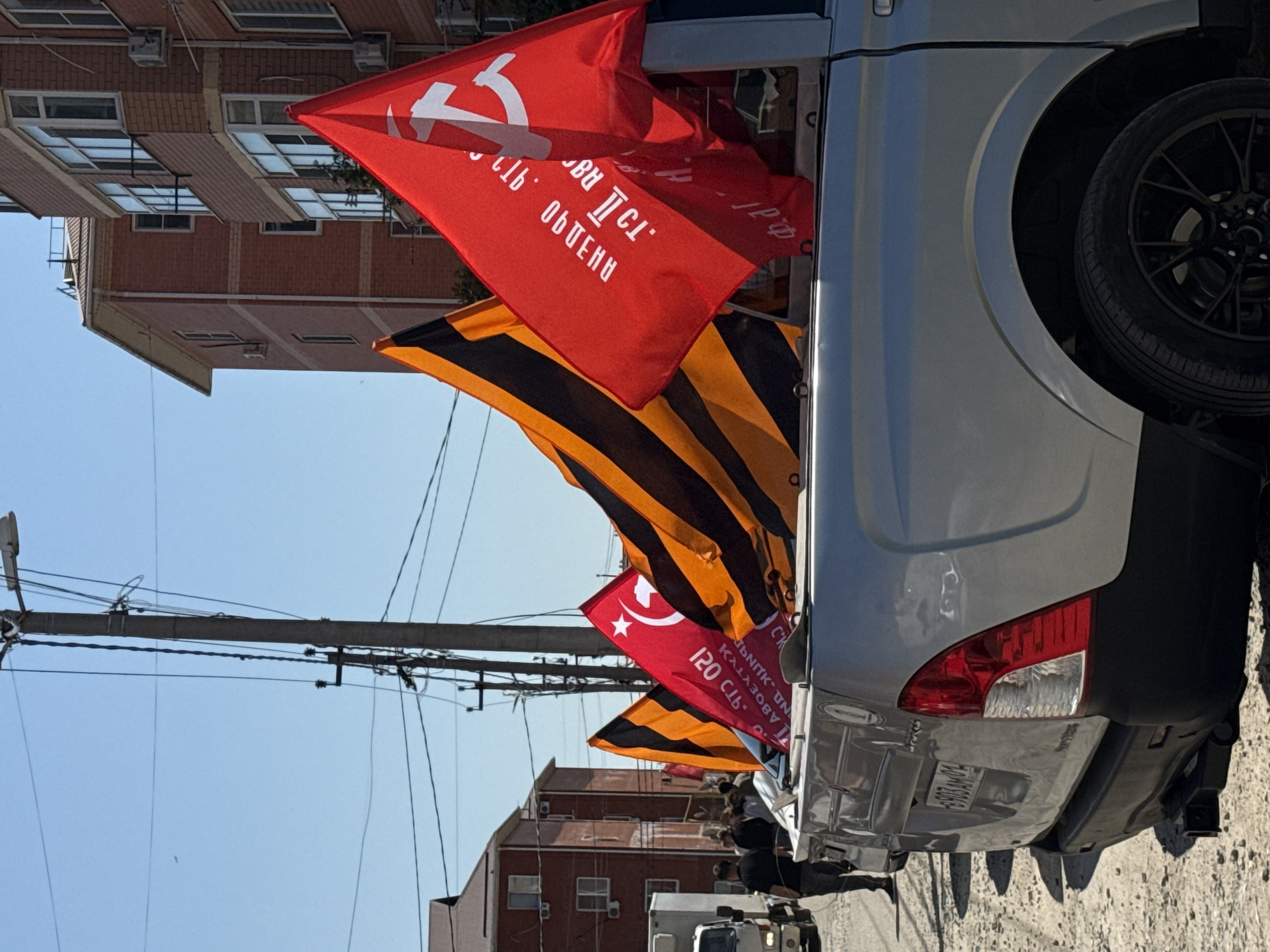The image shows the rear of a gray van parked on a street in an urban area, likely in an unspecified city. The van has a black spare tire mounted on the back. Several flags are displayed, including a red flag with a hammer and sickle symbol and Cyrillic text, as well as a flag with a black and orange striped pattern, reminiscent of the Ribbon of Saint George. There are buildings in the background, and utility poles with overhead wires. A person and what appears to be another vehicle are visible in the distance. The license plate of the gray van is partially visible but unreadable.