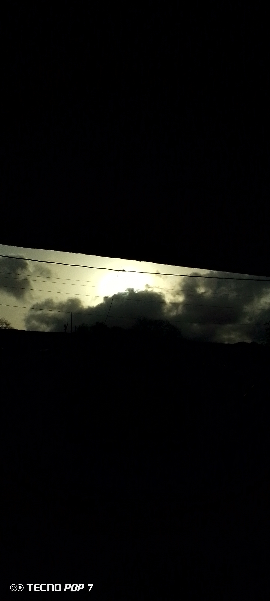 A low-angle, nighttime photograph reveals a dim, overcast sky with a hint of light breaking through the clouds. In the mid-ground, a dark silhouette of trees and possibly a structure form a vague horizon. Power lines stretch across the illuminated space, and the majority of the image is shrouded in darkness. The bottom of the image includes the words "Tecno Pop 7." The precise location and time are unable to be determined.