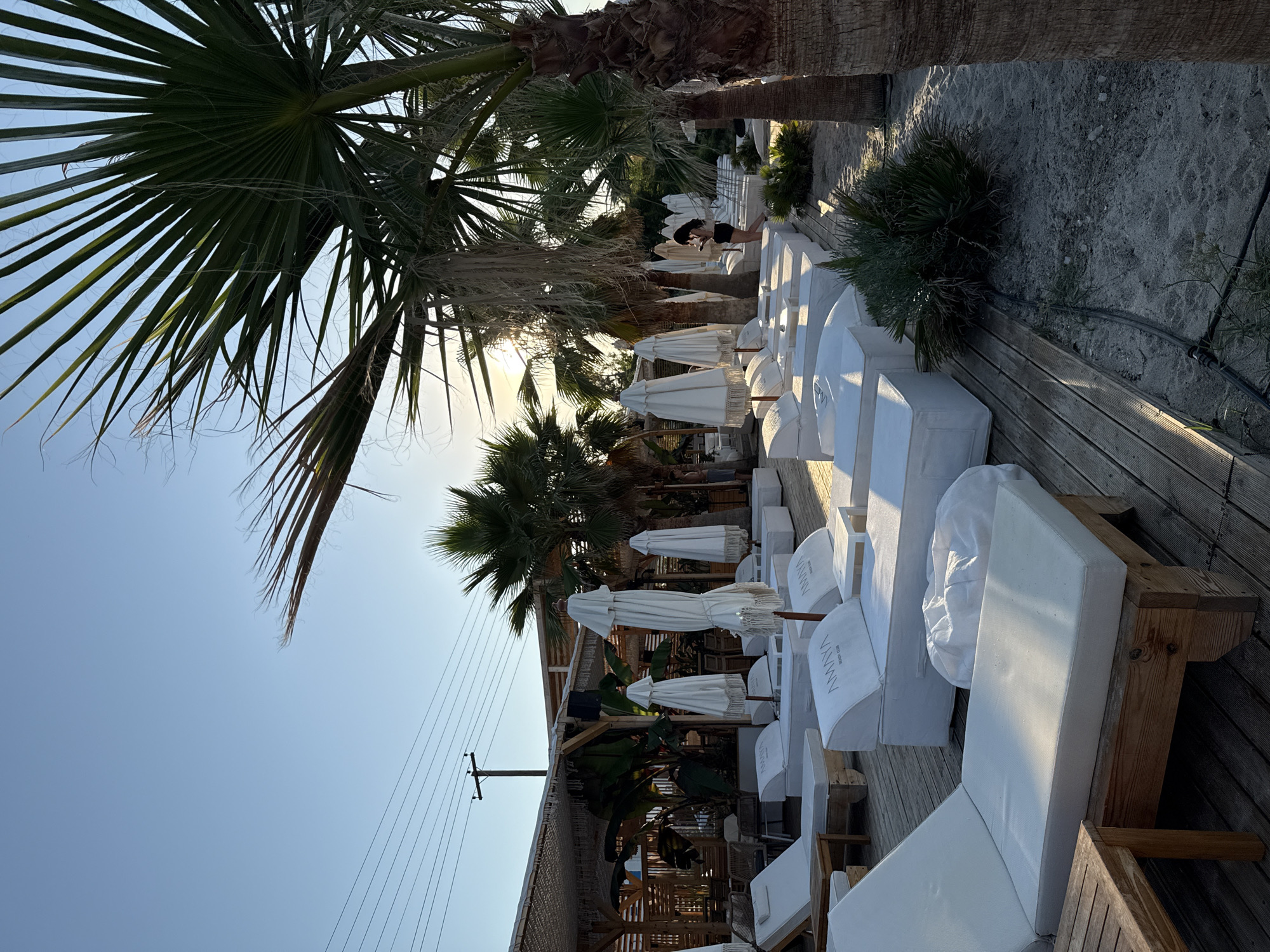 A beach club in Pallini Municipal Unit, Greece, is depicted in this image. The scene is set during daylight with a clear sky and sunlight filtering through palm trees. The beach club features numerous white lounge chairs with umbrellas, arranged on a wooden platform and sand. In the background, a covered seating area with additional chairs and tables is visible, along with tropical plants. One person, a woman with dark hair, is walking amongst the lounge chairs. Another person, a man, is standing near the palm trees on the right side of the image. The overall atmosphere is serene and suggests a relaxing environment for patrons.