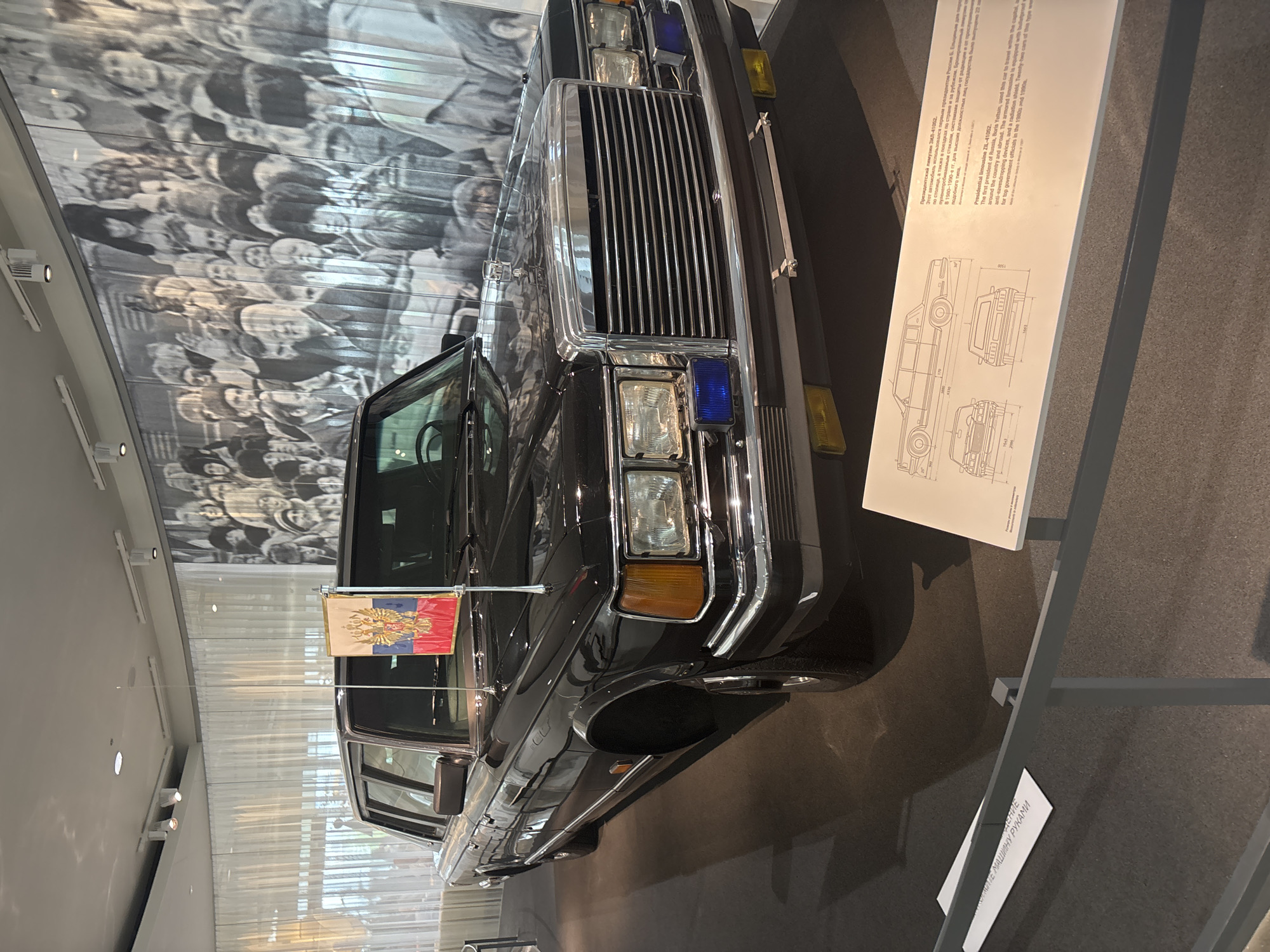 A black ZIL-41052 presidential limousine is centrally displayed indoors at the Boris Yeltsin Presidential Center in Yekaterinburg, Russia. The vehicle, a long-wheelbase sedan, features a prominent chrome grille, rectangular headlights, amber turn signals, and two blue emergency lights integrated below the headlights. A small Russian Federation flag, bearing the national double-headed eagle crest, is affixed to a flagpole on the front right fender. The limousine is highly polished, reflecting the overhead lighting.
The background consists of a large, black-and-white photographic mural depicting a dense crowd of people, many looking forward or upwards, with various expressions. The floor is dark gray, and a gray metal barrier separates the exhibit from the viewing area. Overhead, a white ceiling is fitted with track lighting and recessed fixtures. In the lower right foreground, a white informational plaque provides technical diagrams of the ZIL-41052 and descriptive text in both Russian and English. The English text identifies the vehicle as the "Presidential limousine ZIL-41052," stating it was used by "the first president of Russia, Boris Yeltsin," for domestic and international travel. It also details the car's armored construction, including "bulletproof glass, anti-smashed windows, and a radiation shield," and notes that "twenty-two cars of this type were produced for top government officials" in the 1990s.