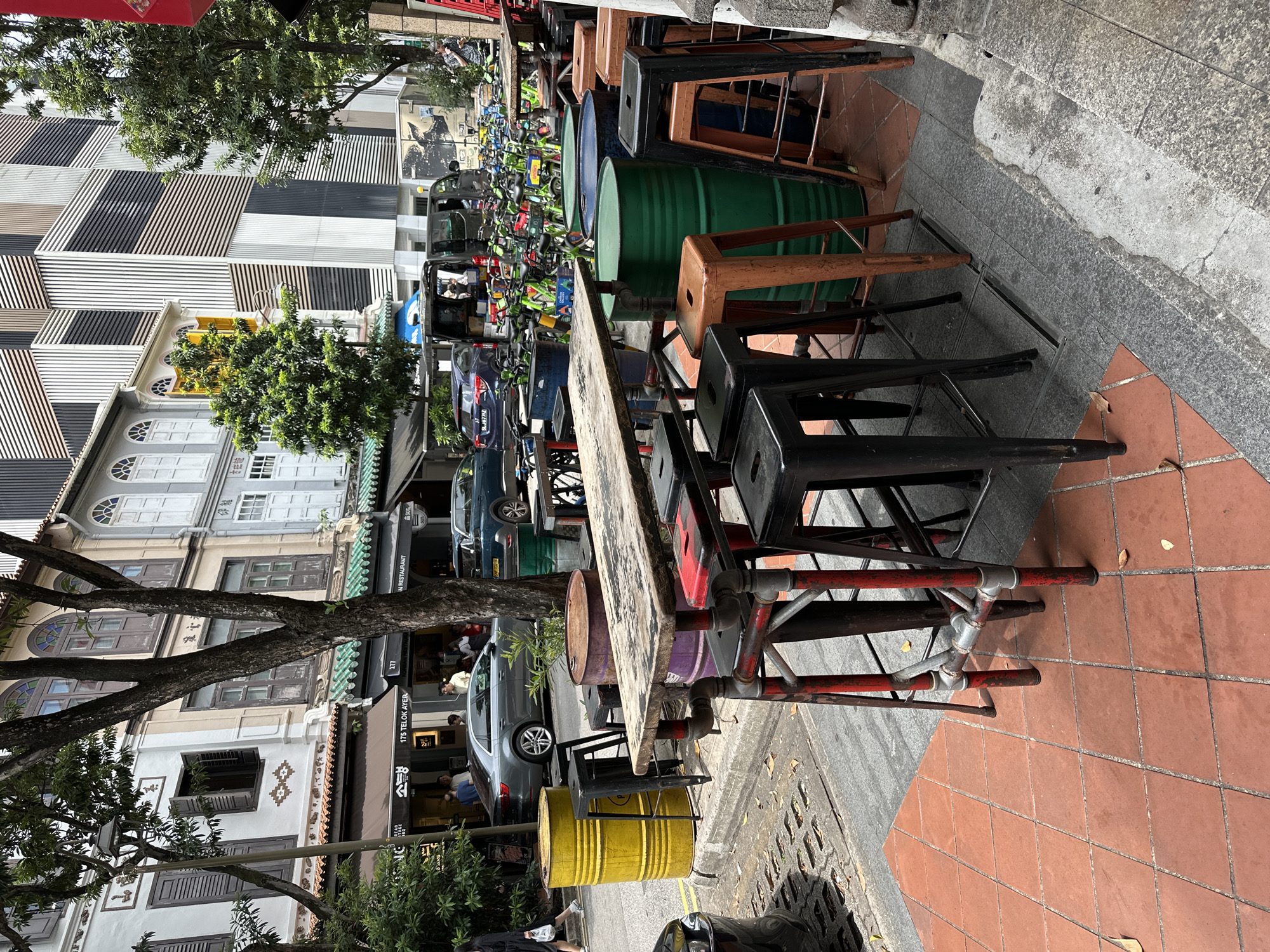 This image depicts an outdoor seating area for a restaurant or cafe located on Telok Ayer Street in Singapore. The scene features a long, rustic wooden table with a metal pipe base, surrounded by various stools. These stools are primarily black metal bar stools and some orange metal stools. To the left of the seating area, there are two large metal barrels, one bright yellow and the other a reddish-brown, serving as potential table bases or decorative elements. Behind the seating, several large green metal barrels are visible, one of which has a dark blue lid.
The background reveals the distinct architecture of shophouses lining the street, characterized by their colorful facades, arched windows, and tiled roofs. Further back, a modern building with a striped facade stands out, suggesting a juxtaposition of old and new urban environments. The street itself is paved with reddish-brown tiles, and a grey concrete gutter runs along the edge. In the distance, a row of bright green rental bicycles is parked.
Several people are present in the scene, though their interactions are not clearly defined. Some individuals are walking on the sidewalk, while others appear to be seated or standing near the establishments. Cars are also visible on the street, including a silver sedan and a dark blue car. The environment is outdoors, under what appears to be daylight, with shadows indicating the sun's position. Trees with lush green foliage are interspersed throughout the scene, adding to the urban greenery.