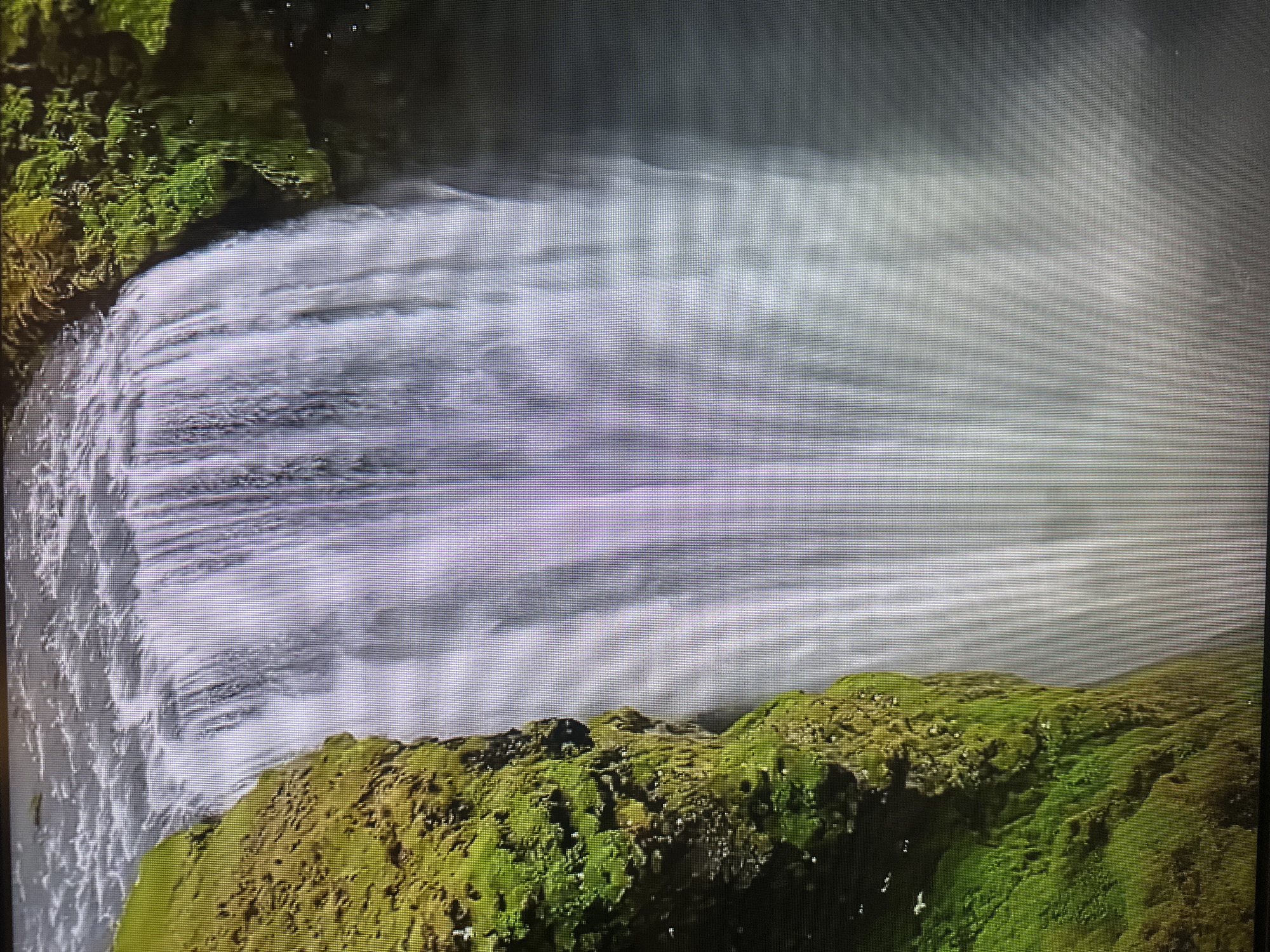 The image shows a close-up view of a powerful waterfall cascading down a moss-covered cliff. The water is white and foamy, creating a mist that blurs the background. The cliff on the left is vibrant green, with textures of moss and rock visible. The image does not depict any elements that would place it in Kyiv, Ukraine; it is a natural landscape scene.