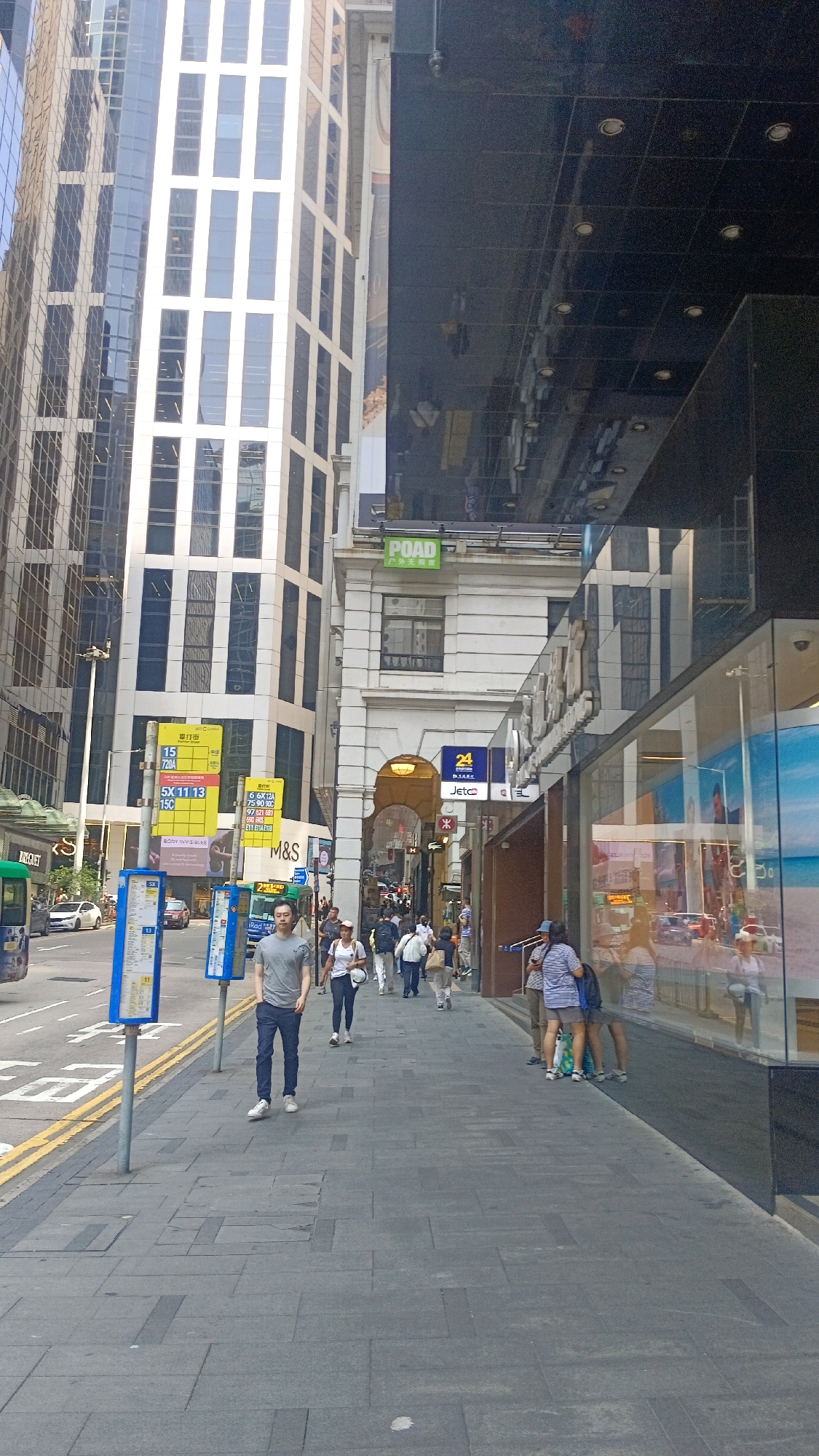 This is a street-level, eye-level, medium shot photograph taken on a sunny day in Hong Kong, China. The perspective is looking down a sidewalk lined with shops and towering modern buildings. On the left side of the image, a tall, glass-fronted building with a grid-like facade dominates the background. Adjacent to it is an older, white building with arched windows and decorative architectural elements. The sidewalk is paved with gray square tiles.
Several people are walking on the sidewalk. In the foreground, a man in a gray t-shirt and dark pants walks towards the viewer. Further down the sidewalk, a woman in a white t-shirt and dark pants walks away from the viewer, followed by several other pedestrians. Some people are entering or exiting shops.
Various signs are visible, including bus route signs, advertising boards, and shop signs. The reflection of the sky and buildings is visible on the polished black exterior of one of the buildings on the right. There are also streetlights and traffic lights.
In the street, cars and a bus are visible. The environment suggests a busy urban commercial district. The overall impression is of a bustling city environment on a clear day.