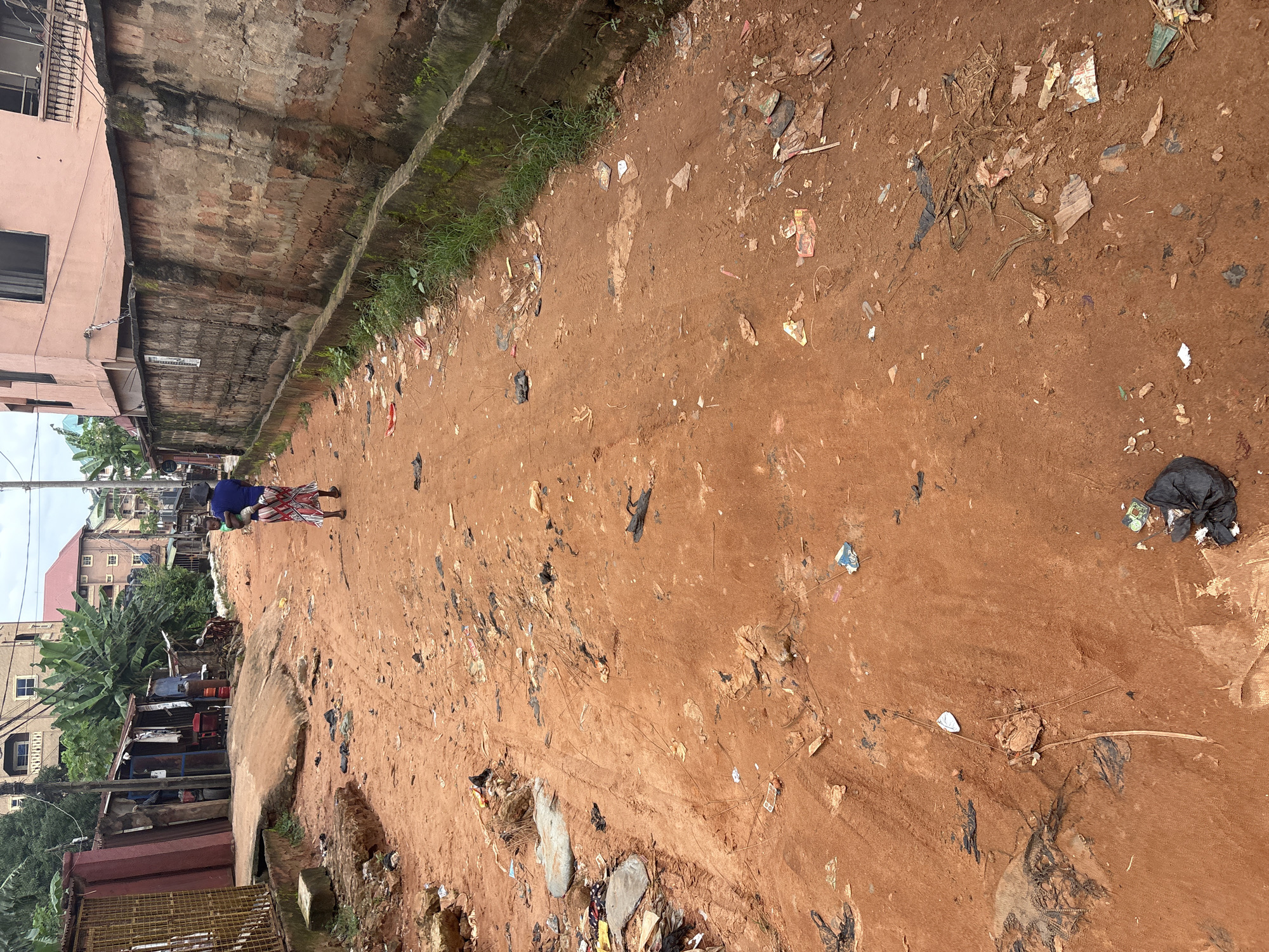 The image depicts a dirt road in Nkpor, Nigeria, strewn with various types of litter, including plastic bags, paper scraps, and other refuse. The road is unpaved and appears to be a common thoroughfare, with tire tracks visible in the reddish-brown dirt. On the left side of the road, there are makeshift stalls or structures with corrugated metal roofs and some visible goods. Beyond these structures, residential buildings are visible. On the right side, a tall, rough-hewn brick wall runs along the road, with some greenery growing at its base. A person, possibly a woman wearing a colorful skirt and blue top, is walking away from the camera, carrying a child on her back. The overall scene suggests a busy, lived-in urban environment with significant waste management challenges.