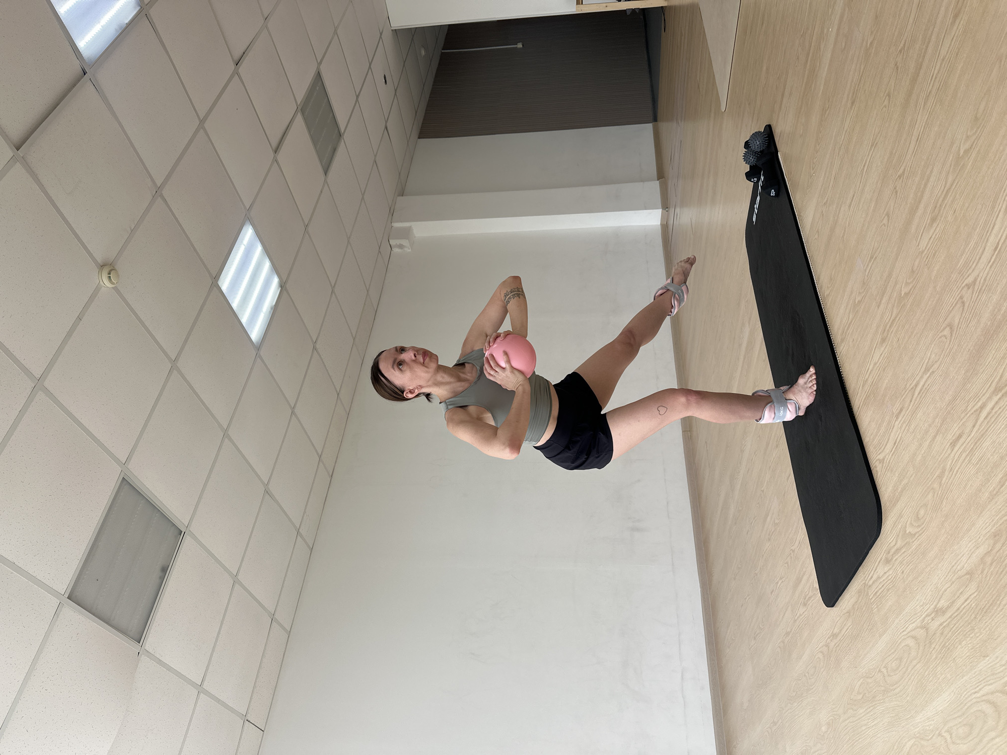 A woman is performing a fitness exercise on a black yoga mat on a wooden floor in a room. She is wearing a gray tank top, black shorts, and weighted ankle cuffs. She is holding a pink ball with both hands in front of her chest. Her left leg is bent and her right leg is extended behind her, with her right foot pointed. The room has white walls and a white drop ceiling with fluorescent lights. There are two small weights and two textured massage balls on the yoga mat behind her. The location is Votkinsk, Russia.