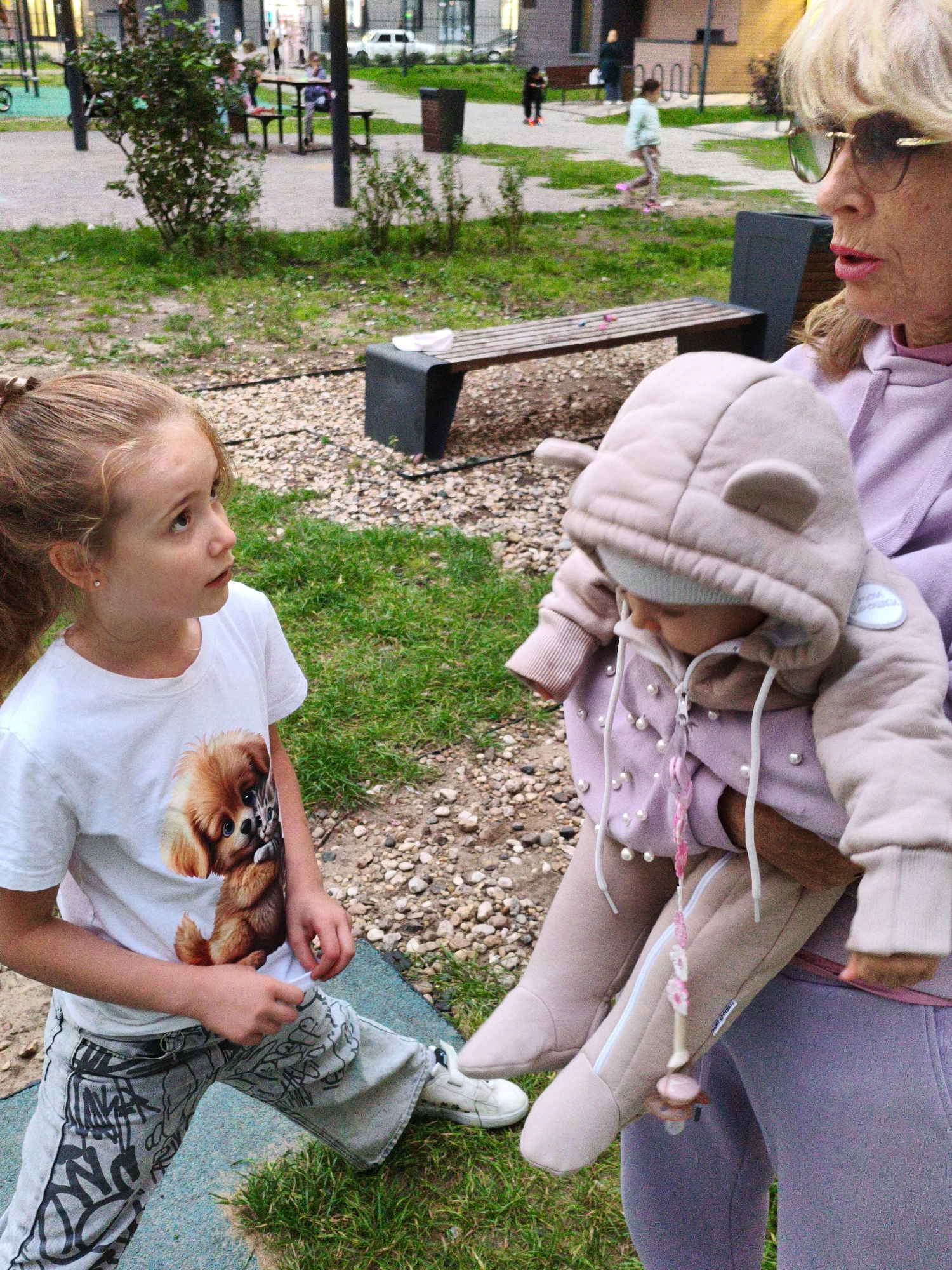 The image depicts a park or playground setting in Lyubertsy, Russia, featuring three individuals. In the foreground, a young girl with auburn hair tied in a ponytail stands looking upwards, wearing a white t-shirt with a graphic of a dog and a cat, and grey patterned pants. To her right, an adult woman, likely a grandmother, holds a baby. The woman has blonde hair and is wearing sunglasses and a pink hoodie. The baby is dressed in a light brown hooded outfit with decorative pearls and a pacifier. The background shows a bench, gravel pathways, grass, and a building in the distance.