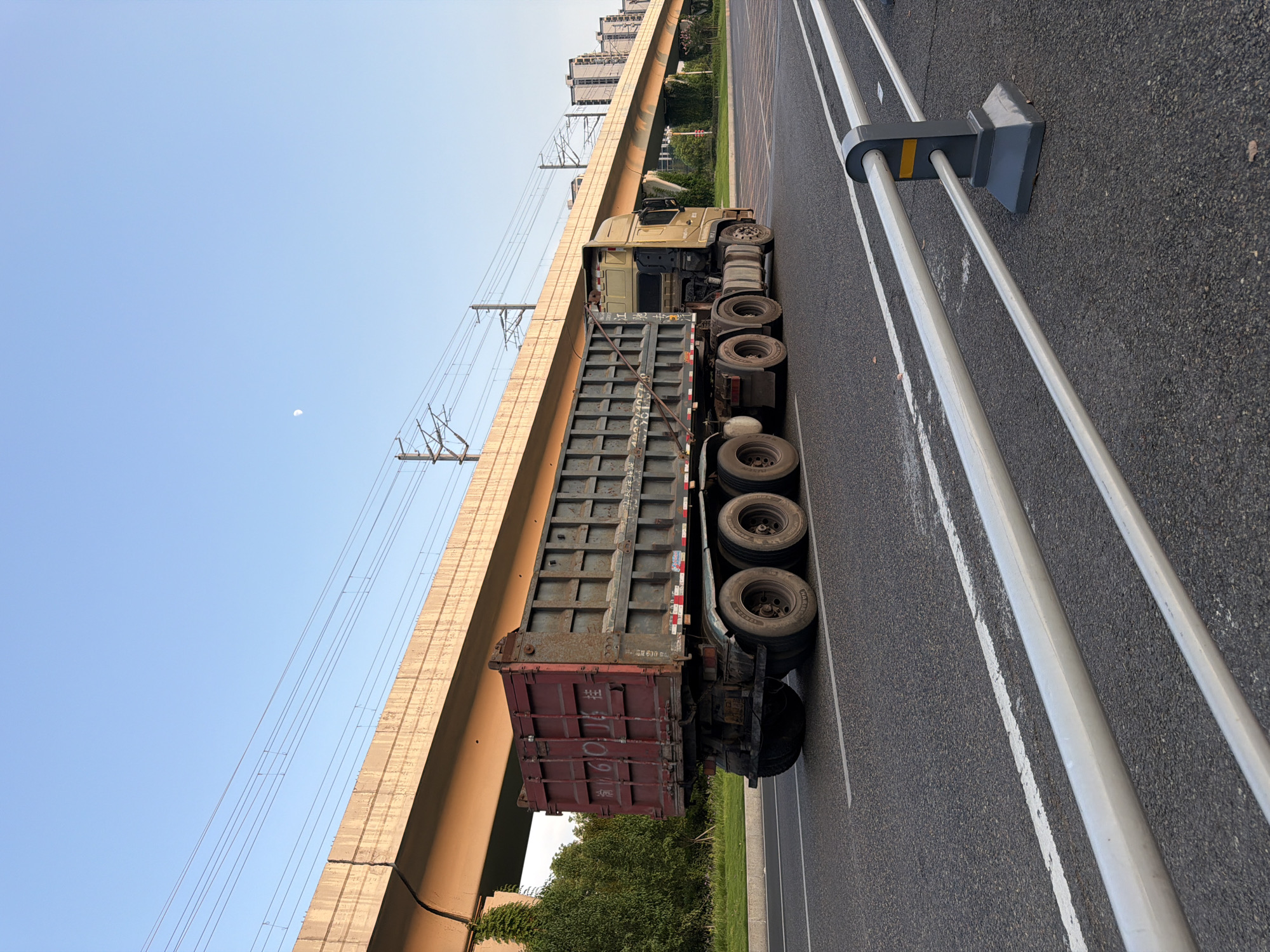A long, multi-axle dump truck with a red and gray trailer travels on a paved road under a golden-colored elevated structure, possibly an overpass or a train track support. The truck's trailer has Chinese characters visible on its rear door, appearing to read "6018 桂." The sky is clear blue with a sliver of the moon visible. Trees and distant buildings line the background. In the foreground, a metal guardrail with gray concrete supports runs along the edge of the road. The scene appears to be set in Jiangjin, China, as indicated by the presence of the Chinese characters.