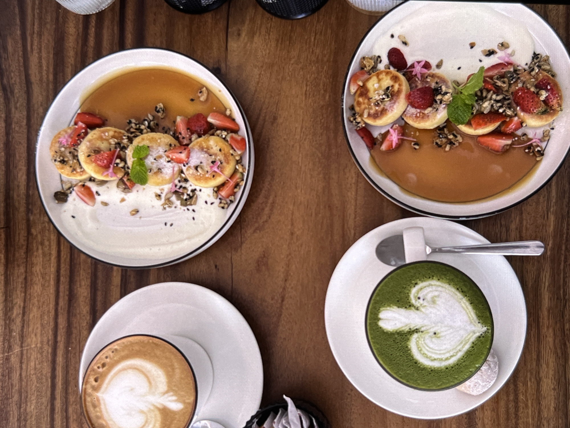 Here is a top-down shot of a wooden table on which are placed two plates of food and two cups of beverages. The plates of food each contain small, round pancakes topped with what appears to be strawberries, edible flowers, and a light sprinkle of seeds or nuts, drizzled with a caramel sauce. One plate has a white sauce beneath the caramel, and the other has what appears to be a white sauce as the dominant presence on the plate. One cup contains a beverage with latte art, and the other has a green liquid, possibly matcha latte, with a similar design. A spoon rests on the plate next to the green drink. A small cube of sugar is visible on this plate. The location is presumed to be Unknown, Indonesia, as this information was provided with the prompt.