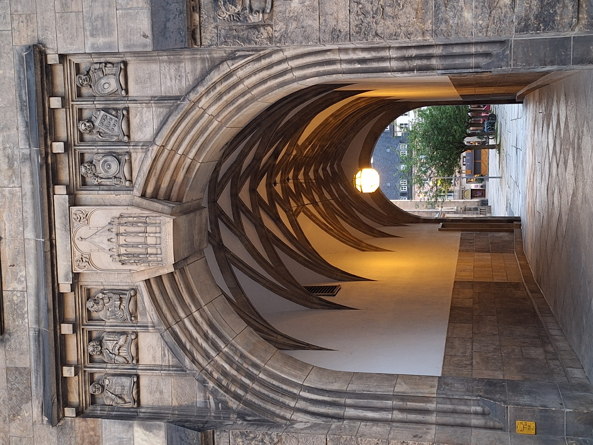 This image depicts an arched passageway, likely a historic architectural feature. The stonework of the arch is intricately carved, with layered, curved elements creating a sense of depth and grandeur. On the left side of the archway, three carved figures in relief are visible, each appearing to hold a shield and helmet, suggesting a theme of defense or historical figures. Above these figures, a more elaborate carved panel depicts a scene that appears to be architectural in nature, possibly a representation of a building or monument.
The passageway itself is illuminated by a warm, yellow light emanating from a spherical lantern hanging from the vaulted ceiling. The interior walls of the passage are smooth and light-colored, contrasting with the textured stone of the arch. The perspective draws the viewer's eye towards the exit of the passageway, where a glimpse of an outdoor scene is visible.
Outside, a tree with lush green foliage stands in what appears to be a cobblestone plaza. Several buildings with dark roofs and uniformly spaced windows can be seen in the background, hinting at an urban setting. The light suggests it could be late afternoon or early evening, with a soft, diffused quality to the illumination. The overall atmosphere is one of historical significance and architectural beauty. A small yellow sign with black text is visible on the right-hand side of the passage near the floor, though the text is too small to be legible. The image is oriented vertically.