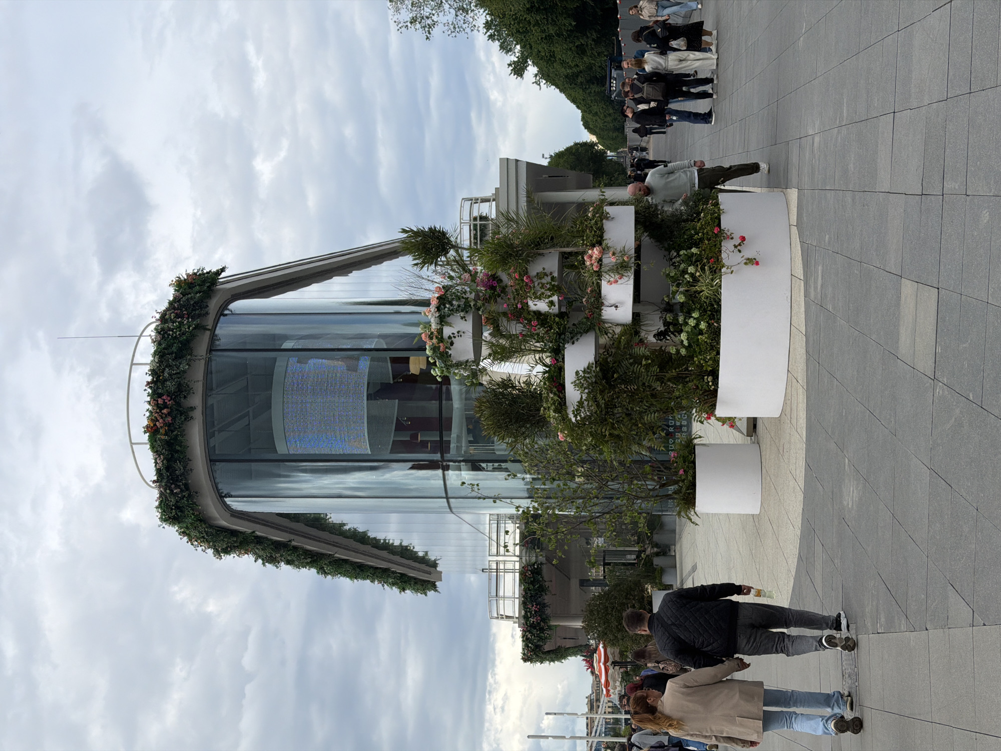 The image captures a scene at Zaryadye Park in Moscow, Russia. The park's distinctive glass-domed pavilion is visible, adorned with vegetation and flowers around its exterior. A digital display can be seen through the glass. The park's paved walkway is visible, populated with people. Some are strolling in groups, while others appear to be waiting in line. To the right of the frame is a series of white planters filled with greenery. The sky overhead is overcast.