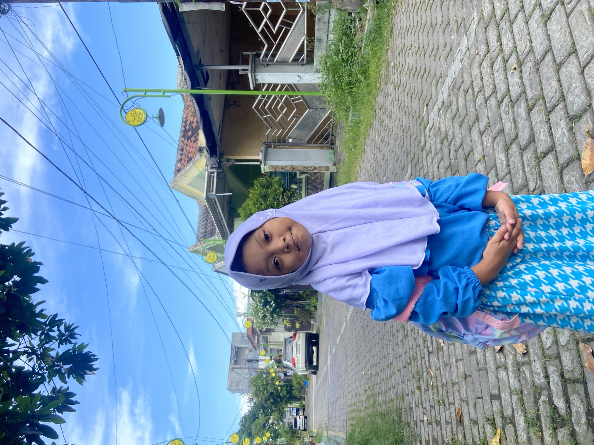 The image, captured in Gelam, Indonesia, presents a bright daytime scene featuring a young girl on a paved street or sidewalk. The girl, the central subject, wears a light purple hijab, a long-sleeved blue top, and a blue and white patterned skirt. A pink strap, possibly from a small backpack, is visible on her left shoulder. She looks directly at the camera with a gentle smile, her hands clasped loosely in front of her.
The background reveals a residential setting with paved surfaces and green patches of grass. Several houses with distinctive architectural elements, including decorative metal gates and terracotta-tiled roofs, line the street. Overhead, a clear blue sky with sparse white clouds is visible, crisscrossed by numerous utility wires. Decorative green streetlights, each featuring a small, circular yellow sign, extend into the distance.
Upon close inspection, the yellow signs bear text in Indonesian. One prominently displays "ES TEH" (Iced Tea) at the top, followed by "MANIS" (Sweet) and additional smaller text, indicating an advertisement for a local beverage. Further down the street, a white car is parked, and other vehicles can be seen in the distance amidst trees and foliage. The overall impression is one of a serene, sunny day in a local neighborhood.