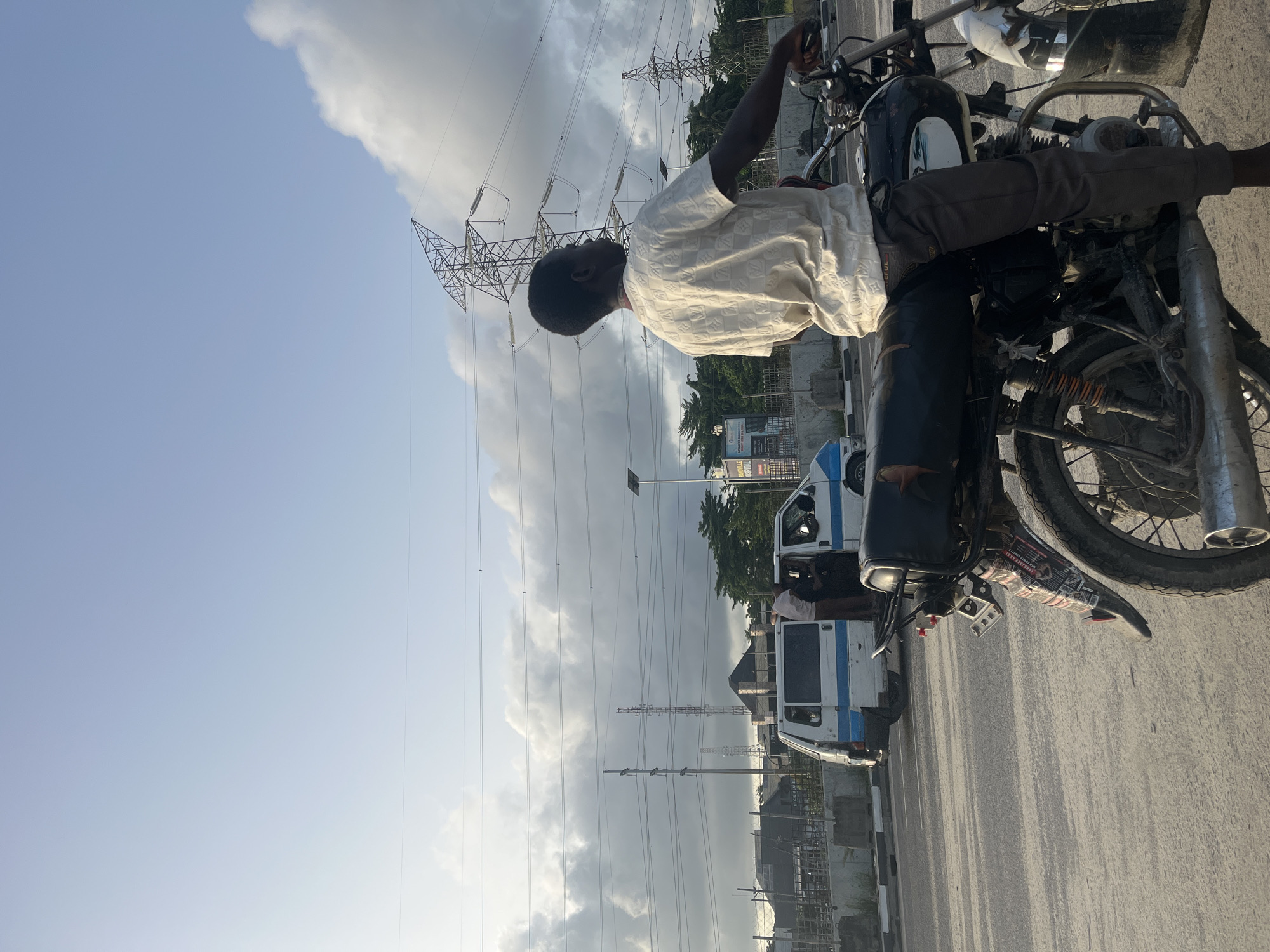 The image depicts a man sitting on a motorcycle in Lekki, Nigeria. The man is dark-skinned, wearing a light-colored, patterned shirt and dark pants. He is positioned on the motorcycle seat with his hands on the handlebars. The motorcycle appears well-used.
In the background, power lines and electricity pylons are visible against a partly cloudy sky. Several white vans with blue stripes are parked along the road. The surrounding area appears to be a typical urban setting in Lekki with buildings and vegetation. The road is paved and shows signs of use.