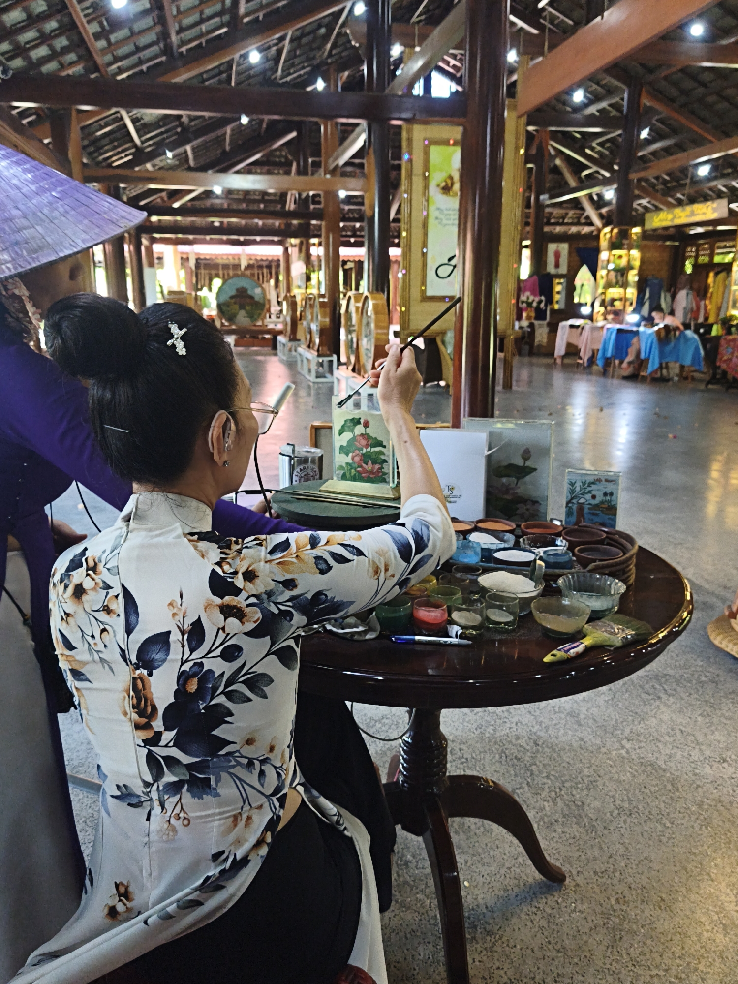 The image depicts an indoor setting, likely a shop or gallery in Nha Trang Ward, Vietnam, characterized by a traditional wooden roof structure and ample lighting. In the foreground, a woman is seated at a round, dark wooden table, engaged in painting. She is wearing a white ao dai with a floral pattern and her hair is styled in a bun with a decorative clip. She wears glasses and appears to have a hearing aid. On the table in front of her are various small bowls containing colored sand, likely for sand painting, along with brushes and what appear to be art supplies. She holds a paintbrush in her right hand, poised over a piece of art on the table. To her left, another individual wearing a purple garment and a conical hat is partially visible. The background reveals a spacious hall with more wooden beams and pillars, display shelves with various items, and other patrons in the distance, suggesting a commercial or cultural establishment.