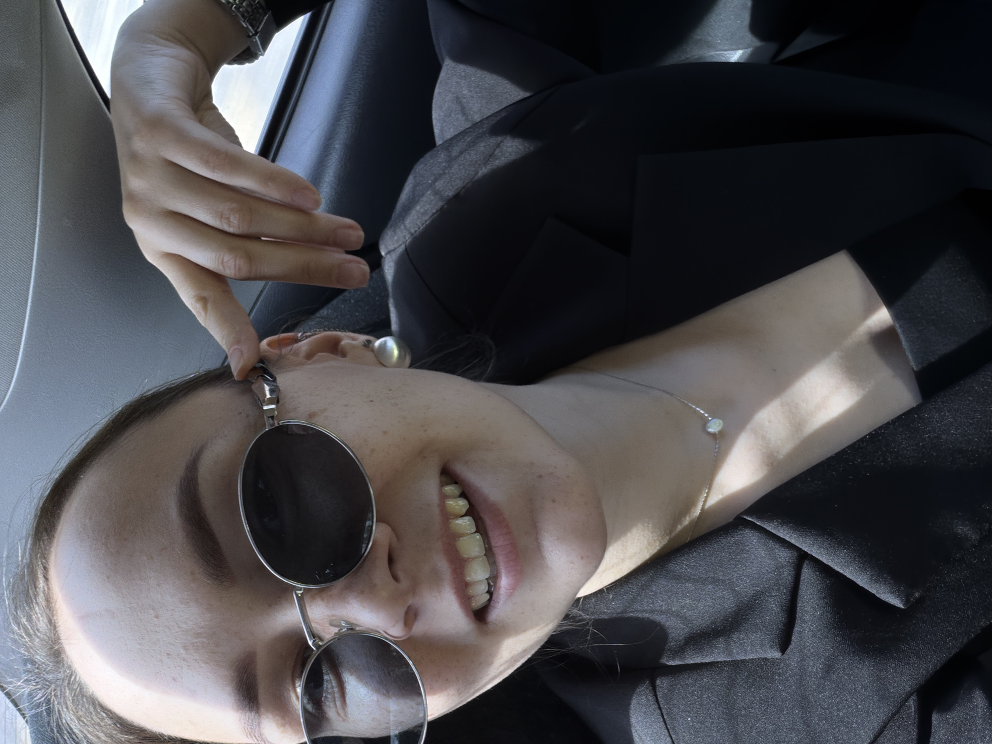 The image shows a young woman with light skin and freckles wearing sunglasses and a black blazer. She is inside a vehicle, likely a car, with the seatbelt visible. Her right hand is resting on the side of her head, near her ear, and she is looking at the camera with a smile. The background suggests an urban environment with natural light. There are no specific landmarks or indicators to pinpoint the location within Chaikovsky, Russia.