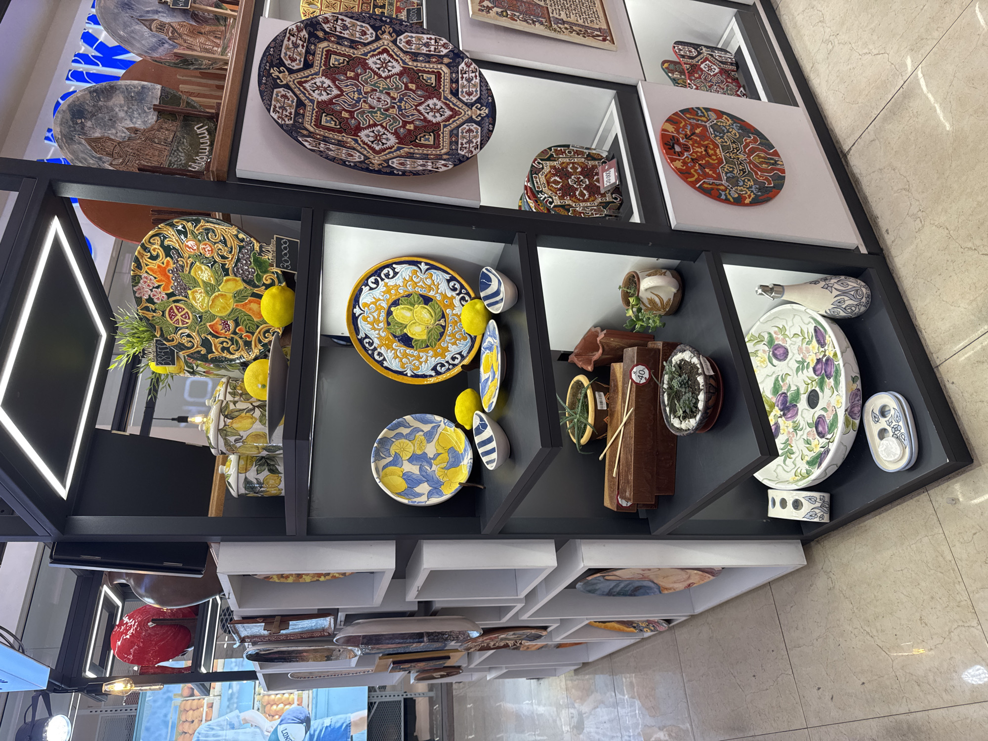 The image depicts a retail display of decorative ceramic items, possibly in a store or market. The items include plates, bowls, jars, and decorative pieces with intricate patterns and designs, many featuring floral and fruit motifs. The display is organized on shelves within a black shelving unit, with some items also placed in white cubby-like compartments on the left. The lighting appears to be artificial, with illuminated strips highlighting the shelves and some overhead lighting. The floor is a light-colored marble or tiled surface.
Based on the visual evidence, specifically the presence of plates with Armenian inscriptions and architectural depictions that resemble Armenian landmarks, this display is likely located in Yerevan, Armenia. The store appears to be a gift shop or a specialty store selling artisanal crafts. There are no people visible in the image. The overall atmosphere is one of a well-lit retail environment showcasing a variety of decorative ceramic products.