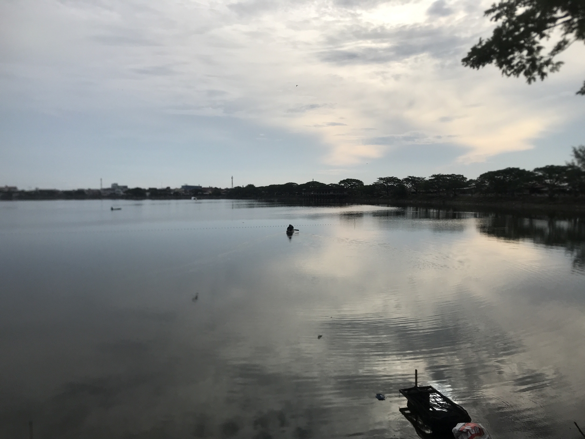 A serene lake scene in Lhokseumawe, Indonesia, captures a tranquil moment at dusk. The sky is a canvas of soft clouds, with hints of pale yellow and blue suggesting the setting sun. The water's surface is calm, reflecting the sky and the distant treeline, creating a mirror-like effect.
In the middle of the lake, a solitary figure is visible in a small boat, paddling gently. The boat and the person are silhouetted against the brighter reflections on the water. Further out, a series of buoys or markers can be seen forming a faint line across the water, suggesting a designated area or perhaps a fishing net.
The background of the scene is dominated by a dense line of trees, which frame the far bank of the lake. Behind the trees, hints of urban development are visible, with buildings and what appears to be a tower or chimney. The overall atmosphere is peaceful and quiet, evoking a sense of calm and solitude. In the foreground, partially submerged in the water, is a dark-colored object that resembles a small raft or a piece of debris, with a plastic bag nearby. The overall lighting suggests it is either early morning or late afternoon. No text is visible in the image.