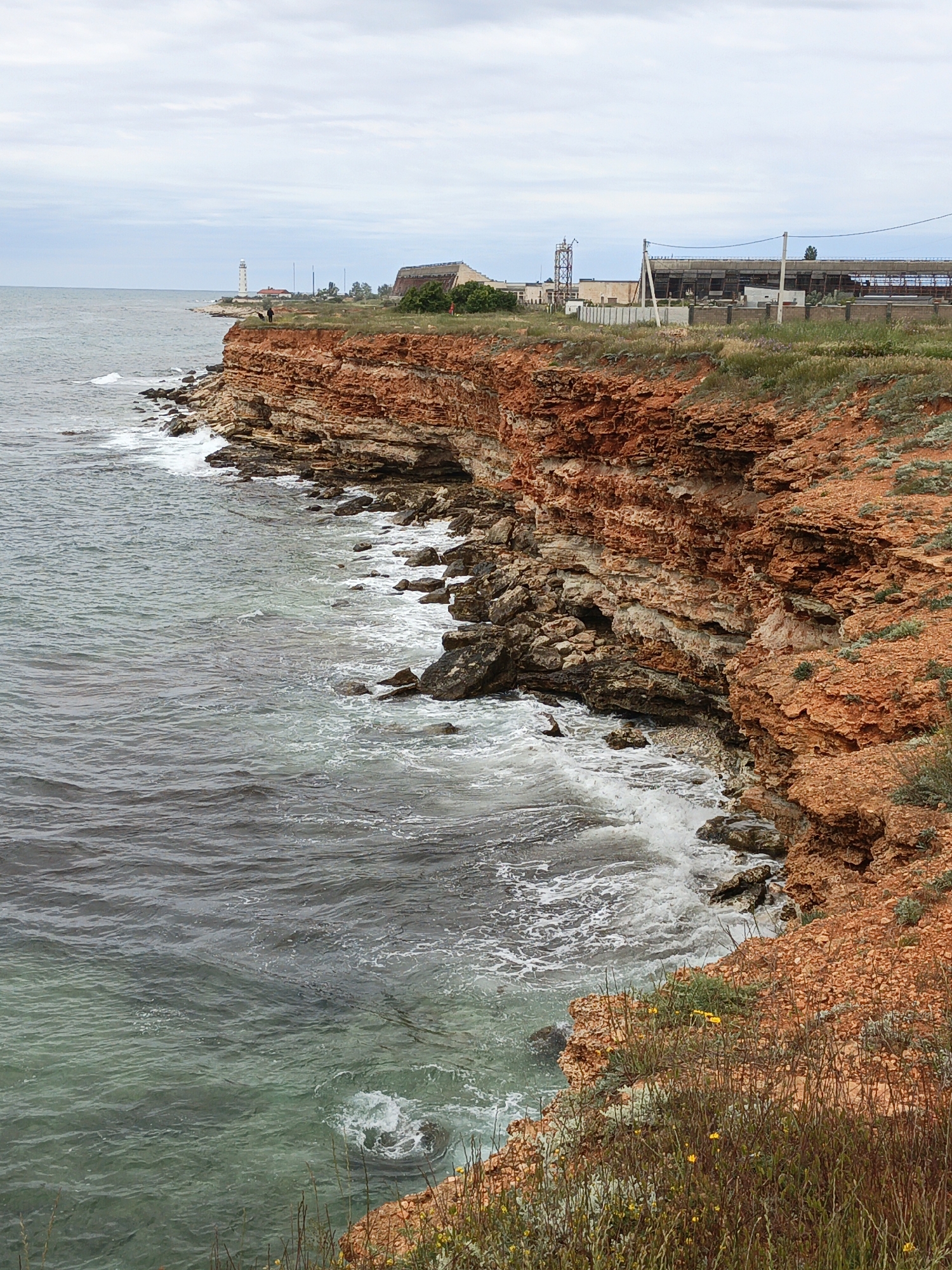 A vertical, medium-shot photograph depicts a layered, red-clay cliff overlooking a body of water. The cliff occupies the right side of the image and recedes diagonally into the background on the left. The top of the cliff is covered with green grass and sparse vegetation. At the edge of the cliff, dried grass and small yellow flowers are visible. The cliff face is red and appears weathered, with some darker rock visible near the waterline.
The water, which fills the left half of the frame, is murky green with white waves crashing against the rocks at the base of the cliff. In the distance, on the left horizon, a lighthouse is visible, as are several buildings and industrial structures on the cliff top.
The location is an unidentified coastal region within the city of Unknown, Unknown.