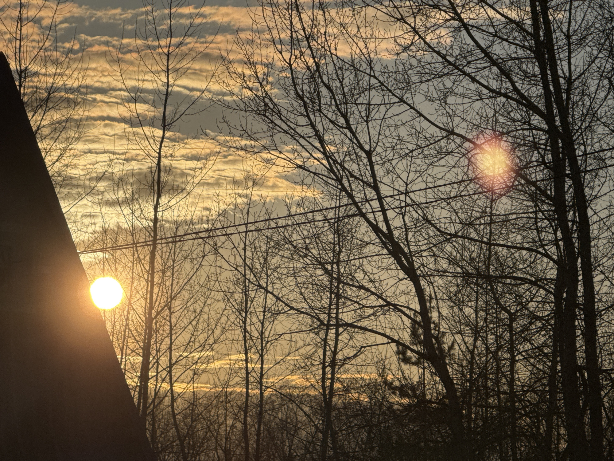 The image captures a sunset scene near Chaikovsky, Russia. Silhouette trees and bare branches dominate the foreground. Overhead, power lines are visible. The sunset is diffused by thin clouds, creating a bright glow in the sky. Part of a roof is visible in the lower left corner of the image.
