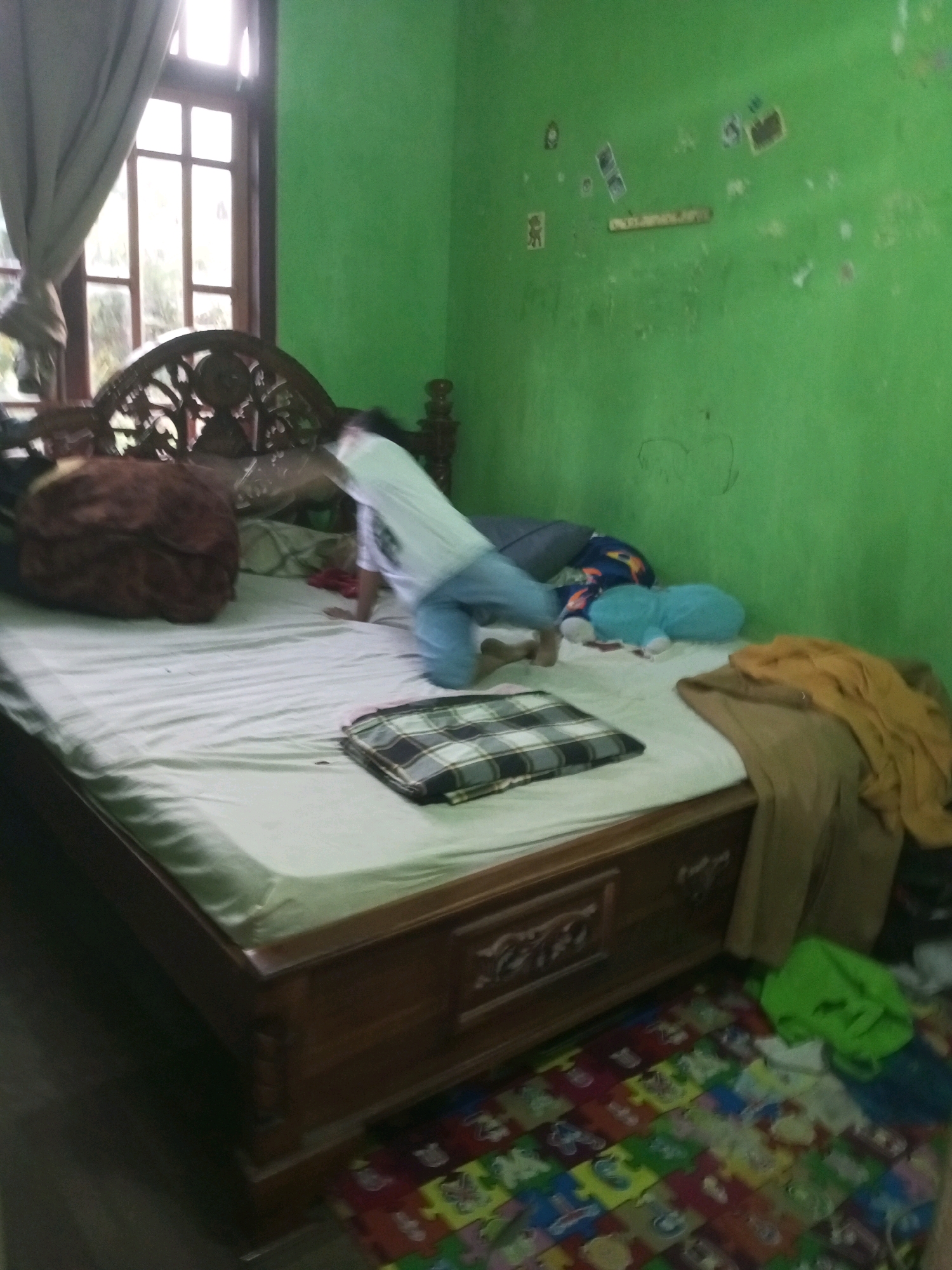 This image depicts an indoor scene within a bedroom in Karangsari, Indonesia. A person, appearing to be a child or young adult, is captured in motion, leaning forward on a bed. They are wearing a white t-shirt and light-colored pants. The bed itself is a prominent feature, with a wooden frame and a white mattress. Various items are scattered on and around the bed, including a large brown fuzzy object, a folded checkered blanket, blue and light blue stuffed toys, and a pile of tan and yellow fabric. The room has bright green walls, some of which have a few faded stickers or drawings. A large window with a curtain is visible to the left, and a colorful puzzle mat lies on the floor to the right of the bed. The overall impression is of a lived-in, informal domestic space.