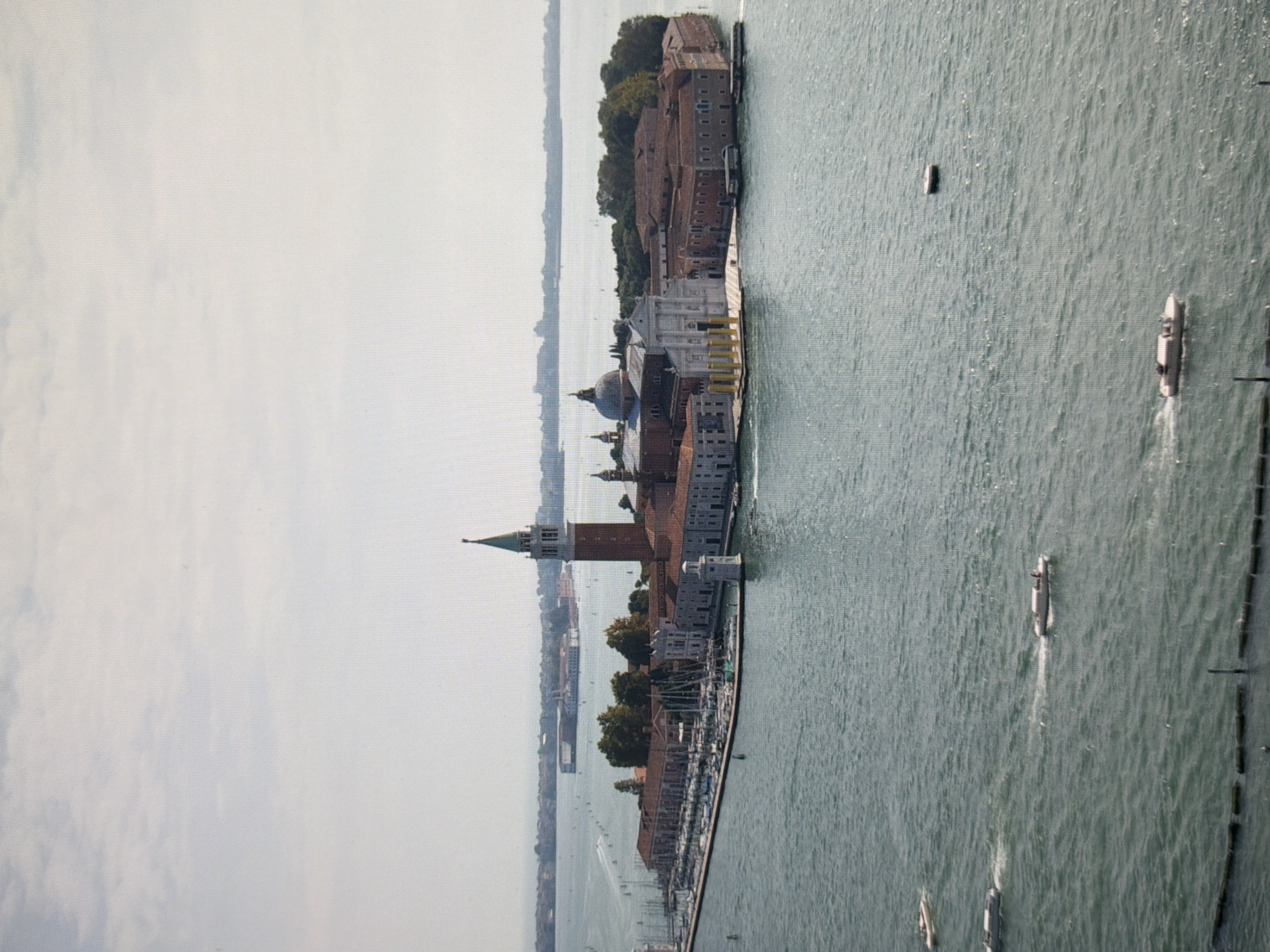 The image depicts an aerial view of the island of San Giorgio Maggiore in Venice, Italy. The island features a prominent brick bell tower with a green spire, a large domed church, and several other buildings with red-tiled roofs. The structures are situated on a green, tree-covered island. The surrounding area is a body of water, likely the Venetian Lagoon, with several boats visible. Some of the boats are moving, creating wakes on the water, while others appear to be docked. In the background, other islands and landmasses with buildings are visible. The sky is a pale blue with scattered clouds. There are no people clearly visible in the image. The scene suggests a sunny day with calm water conditions.