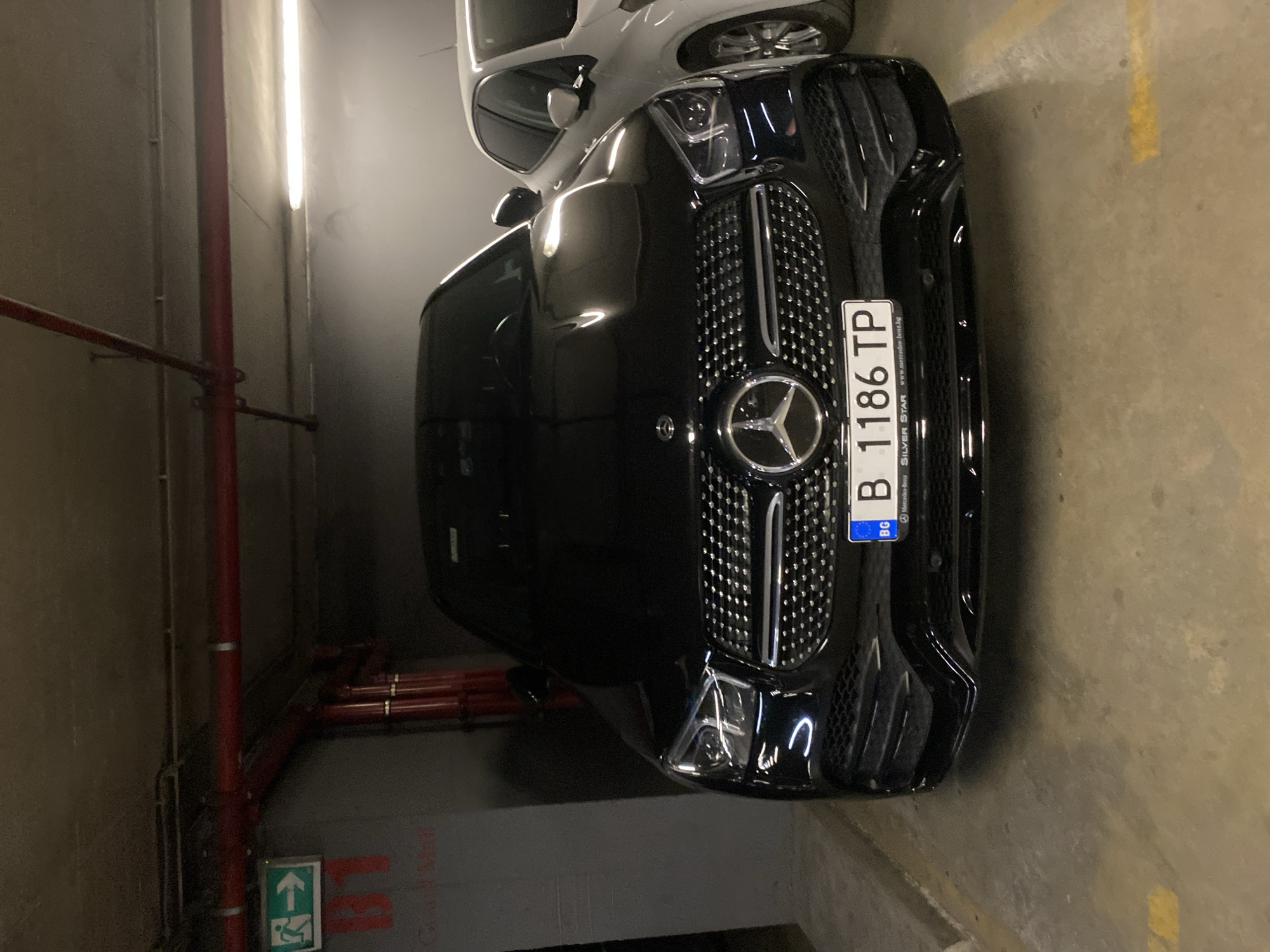 The image shows the front of a black Mercedes-Benz car parked indoors, likely in a garage or parking lot. The car is the primary subject, with its grille, headlights, and license plate clearly visible. The license plate reads "B 1186 TP" and has "BG" on the left side, indicating it is registered in Bulgaria. Below the license plate, "SILVER STAR www.mercedes-benz.bg" is visible. The car is parked on a concrete floor, and a yellow line is faintly visible on the floor to the right of the car. In the background, a white car is partially visible above the black car. To the left of the black car, there are red pipes and a vertical fluorescent light fixture running along a textured wall. An illuminated green emergency exit sign with an upward arrow and a person symbol is visible on the wall in the lower left corner of the image. The lighting suggests an indoor environment, possibly at night or in a dimly lit area. There are no people visible in the image.